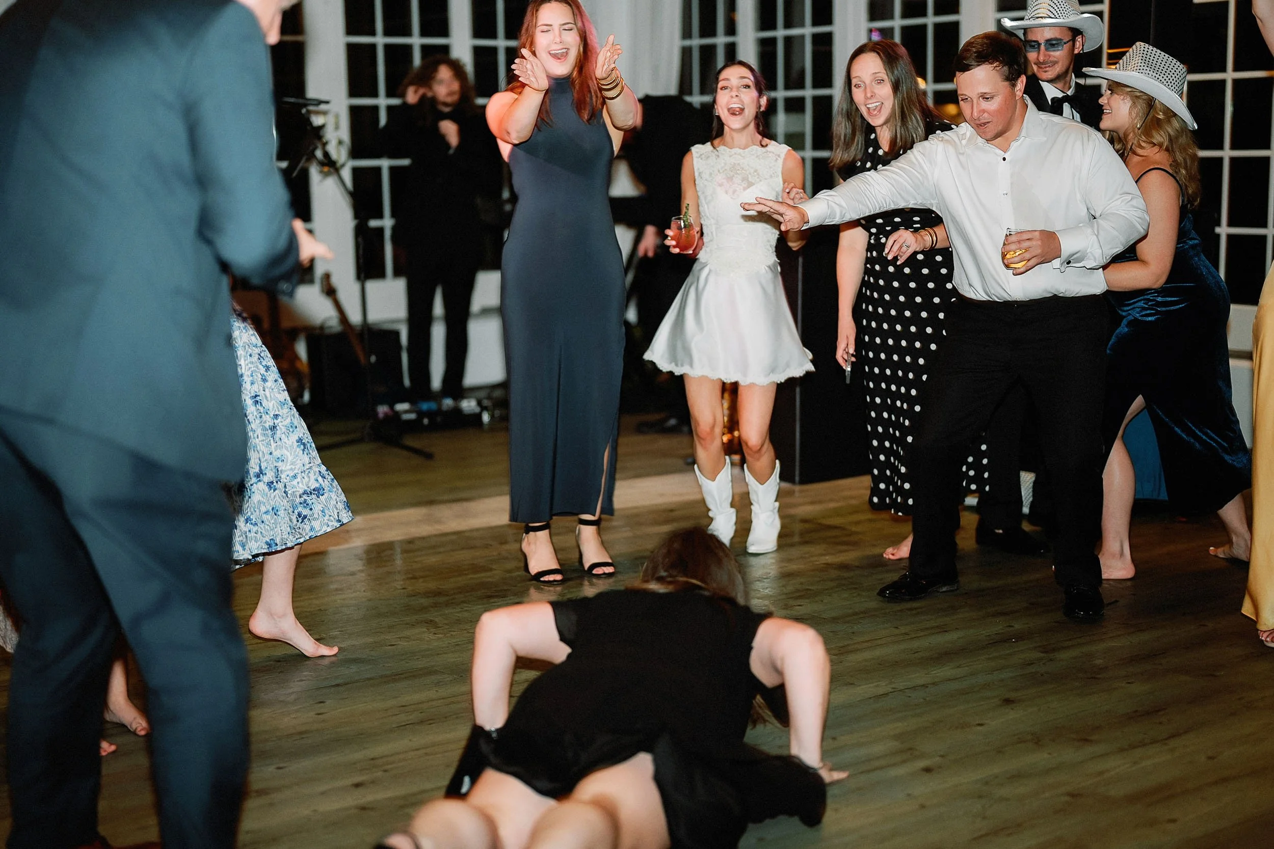 Group of people dancing at a party or celebration, some wearing formal attire and hats, others clapping and cheering, with one person on the floor doing a dance move.