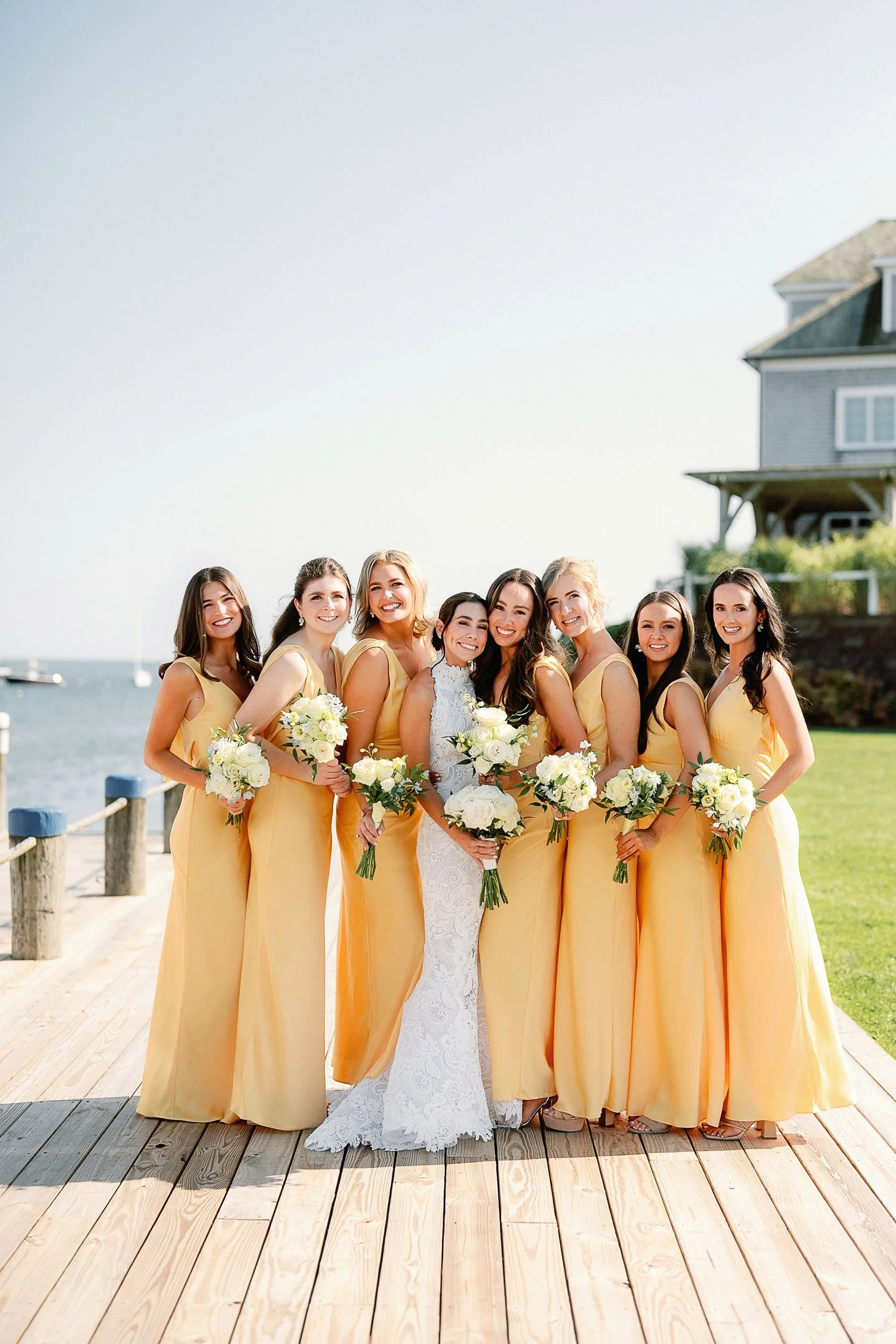 A bride in a white lace wedding dress standing with eight bridesmaids in yellow dresses holding white bouquets by a waterfront on a wooden dock.