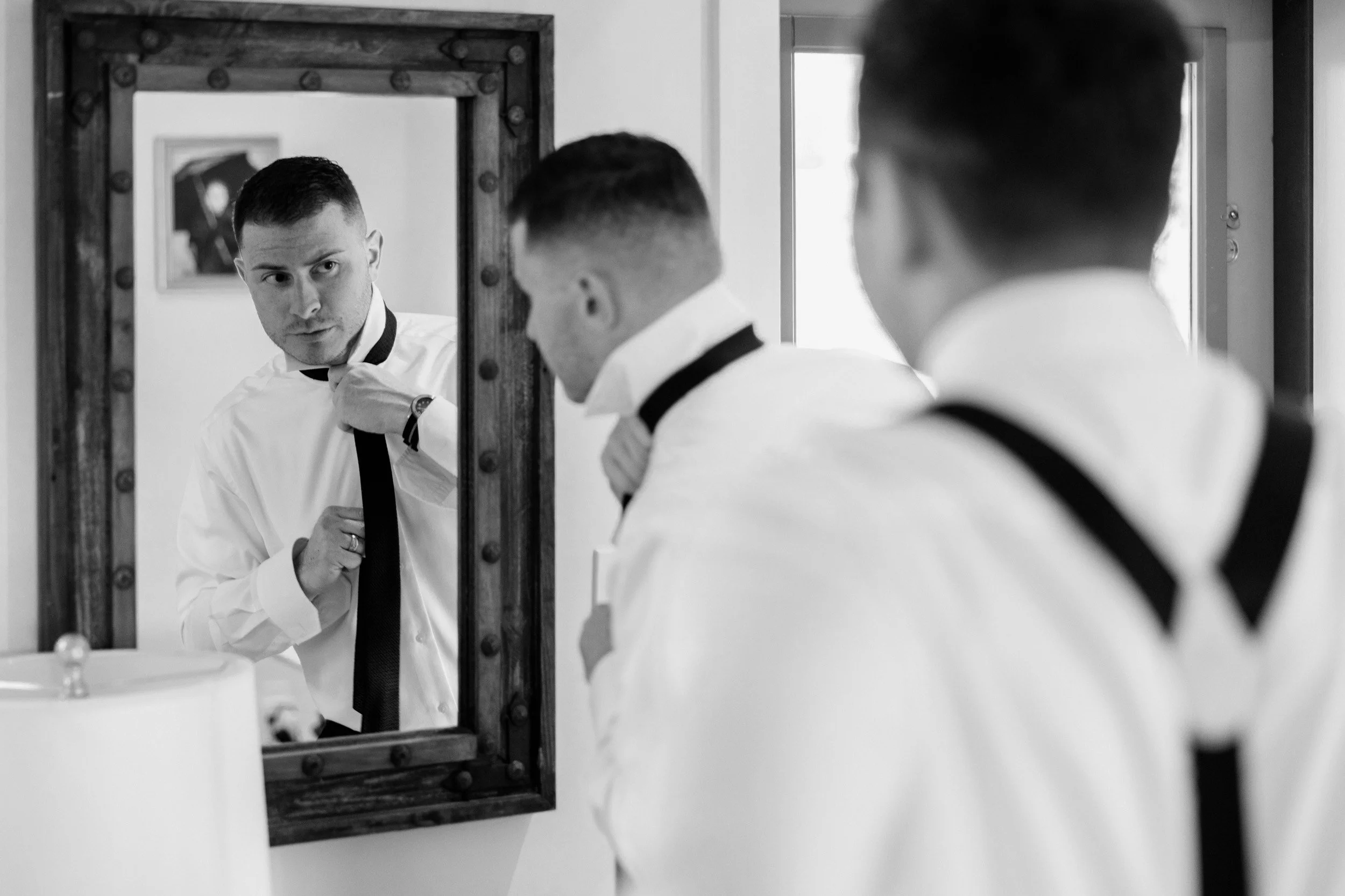 Man adjusting his black tie while looking in a mirror; he is wearing a white shirt and has two other individuals in white shirts with black suspenders nearby.