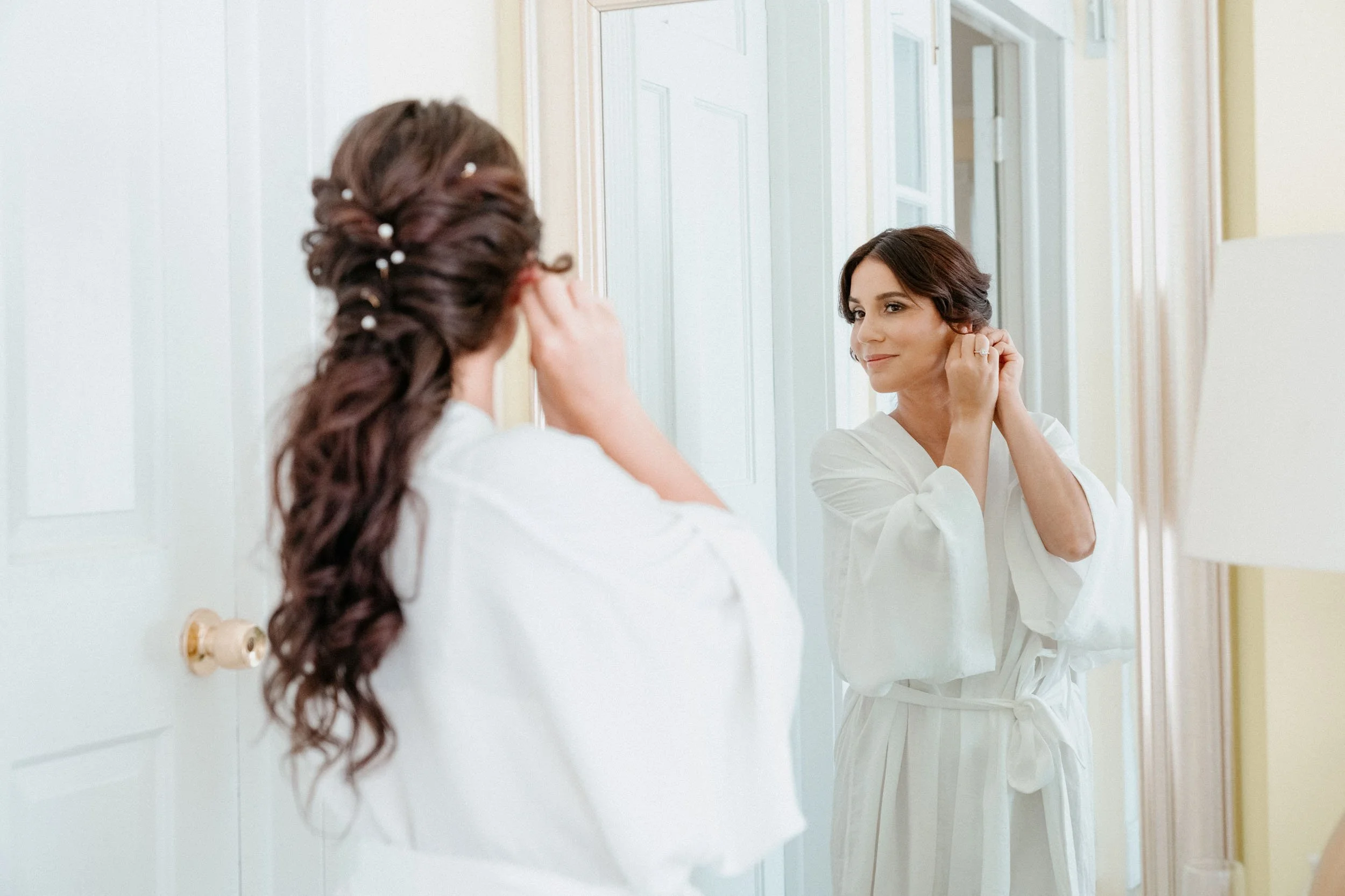 A woman in a white robe is looking at herself in a mirror while adjusting her earring; her hair is styled with decorative pins.
