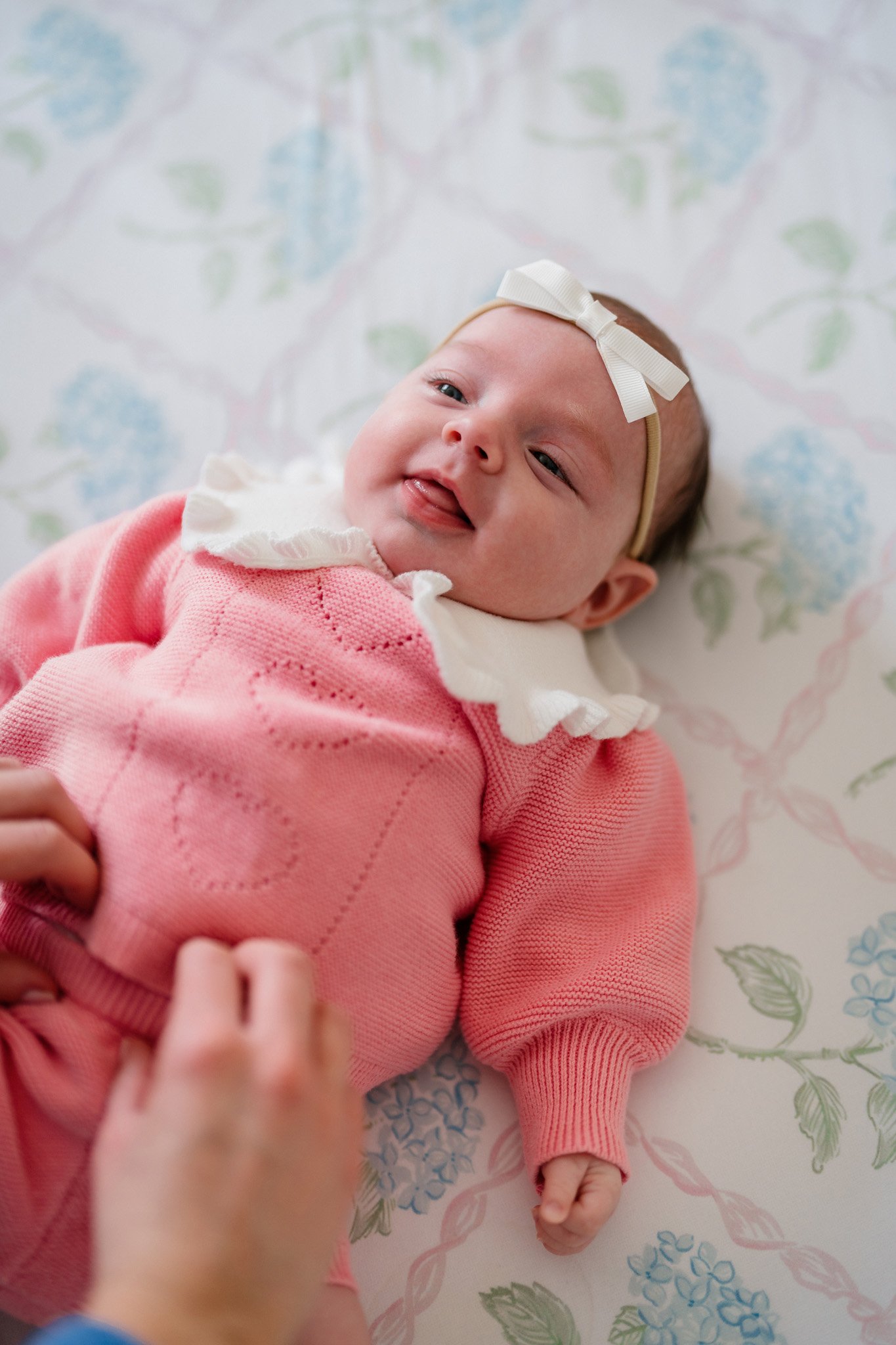 A baby girl lying on a floral patterned sheet, wearing a pink outfit with a white ruffled collar and a white headband with a bow. She is smiling with her tongue slightly sticking out.