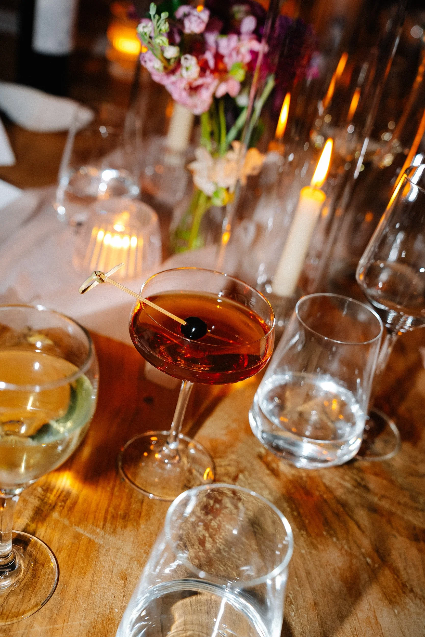 A glass of red cocktail with a cherry garnish, surrounded by empty glasses, on a wooden table with candles and flowers in the background.
