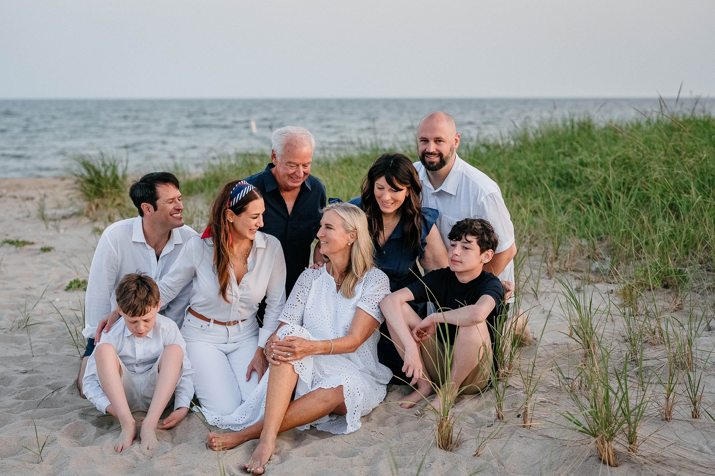 Family group photo on the beach with ocean and grassy dunes in the background, all smiling and interacting.