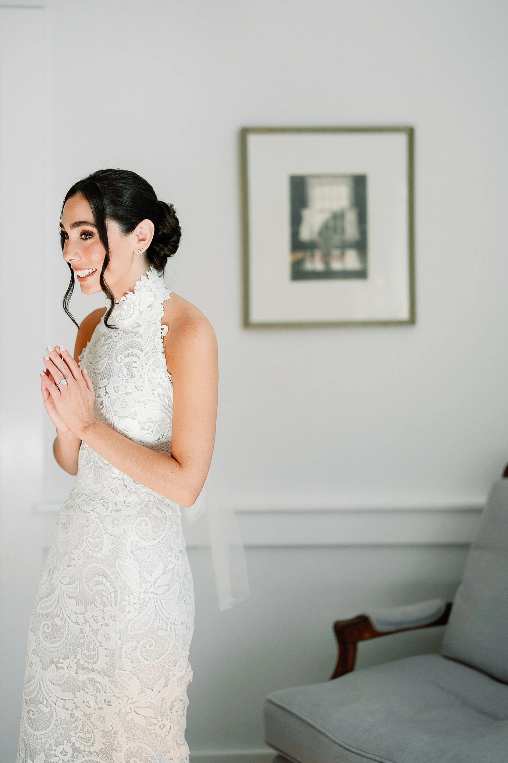 A woman in a white lace wedding dress with her hands clasped, smiling excitedly.