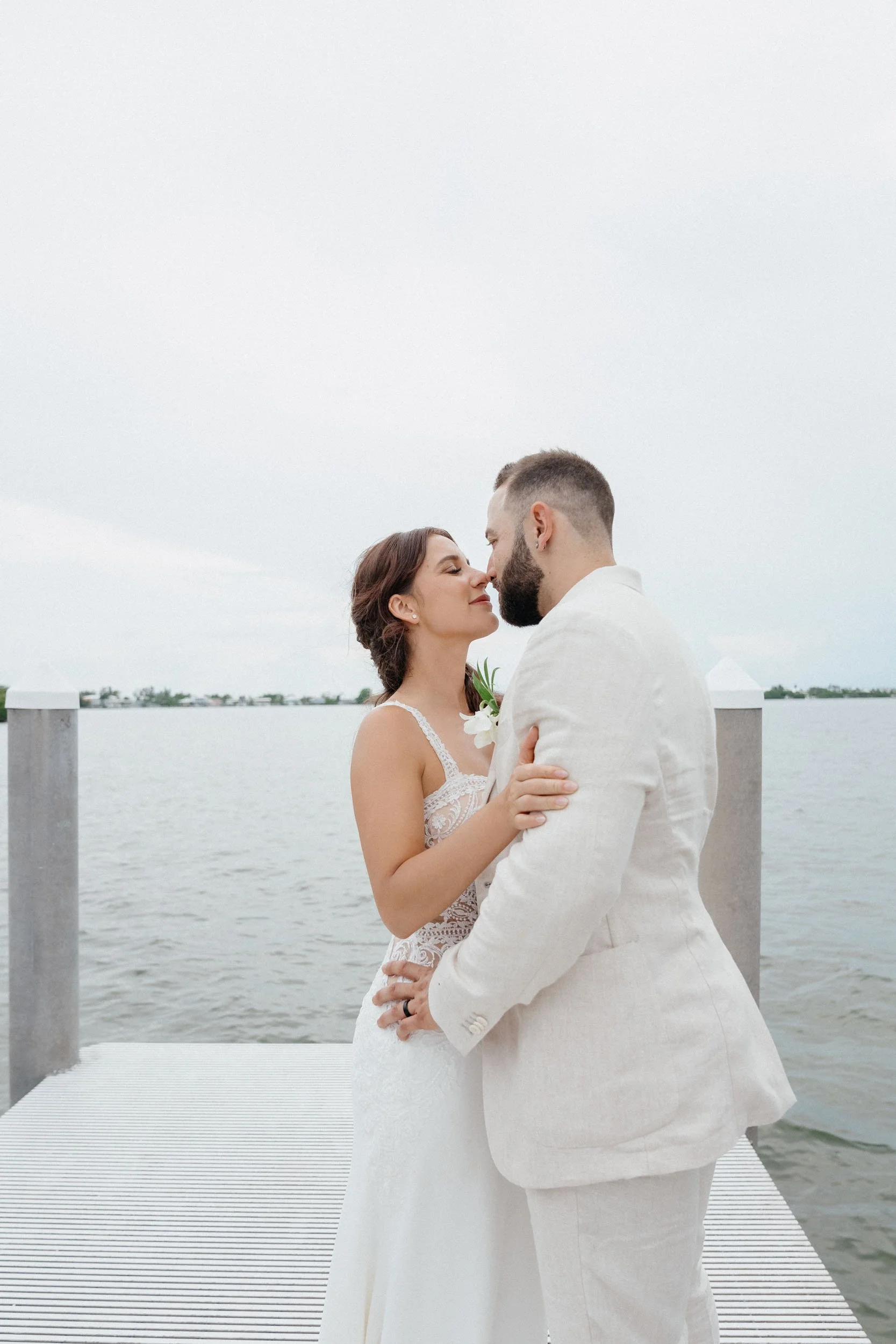 A bride and groom stand closely together on a dock by the water, leaning in for a kiss on a cloudy day.