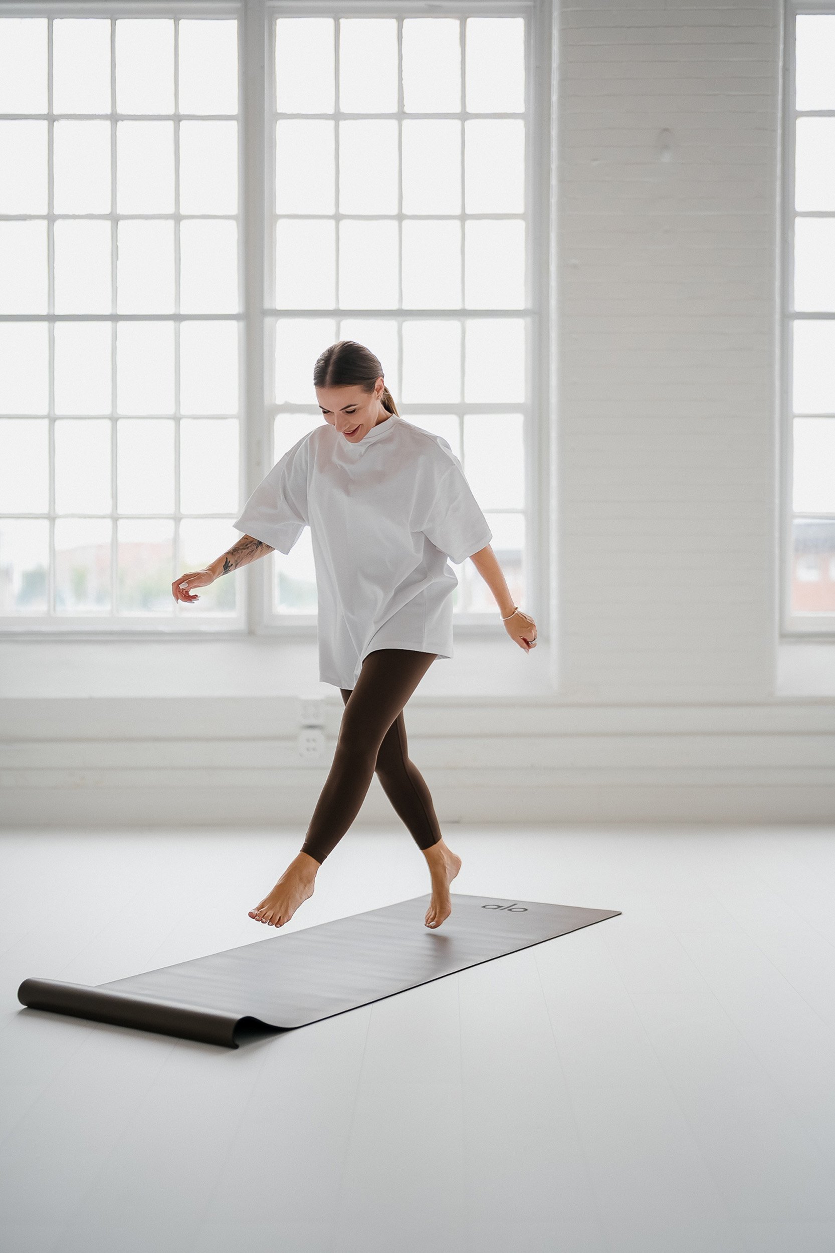 A woman with tattoos on her left arm is standing on a yoga mat in a bright, spacious room with large windows, and she is in the process of stepping or dancing on the mat.