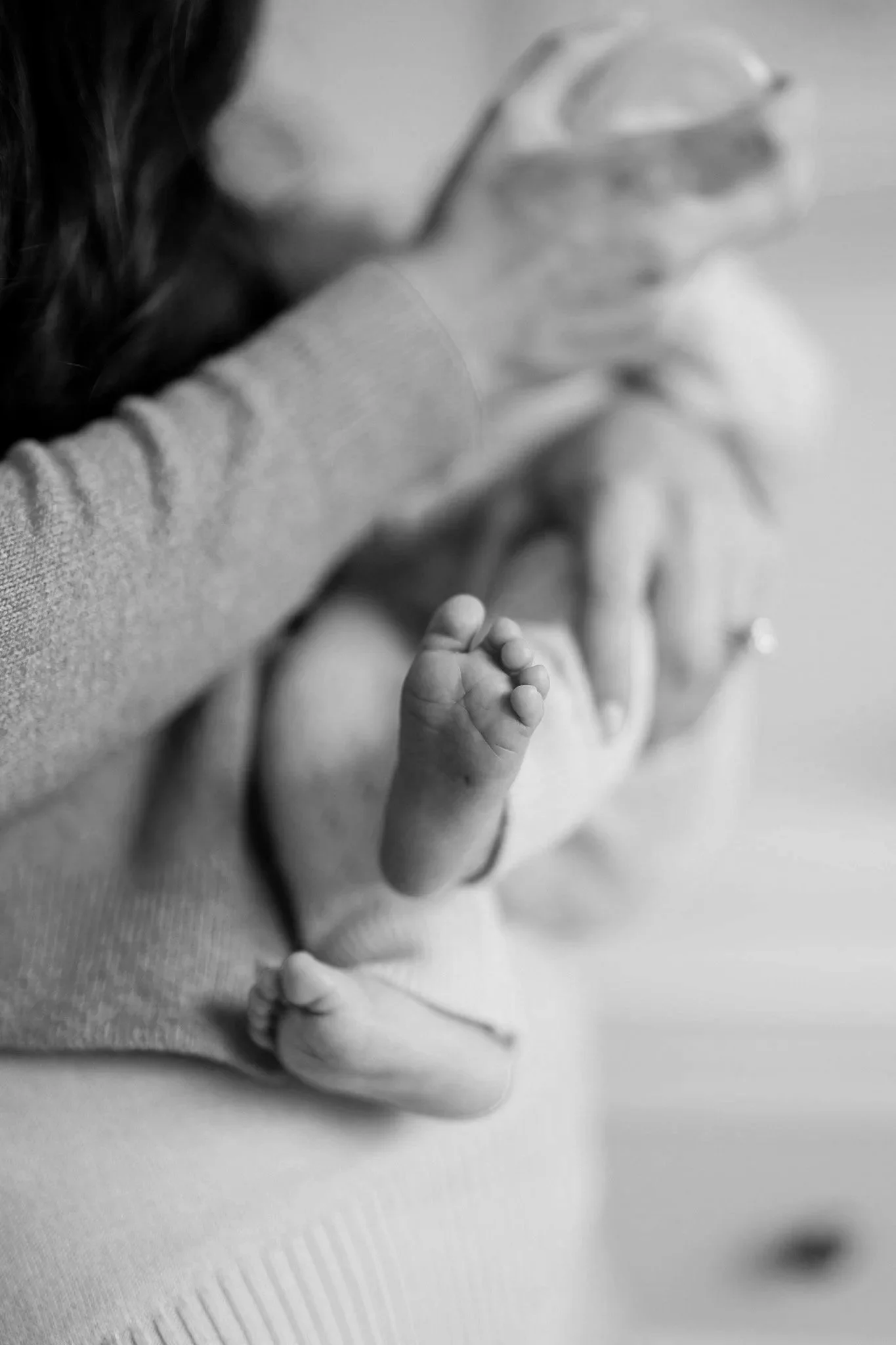 Close-up black-and-white photo of a newborn baby's tiny feet and hand with an adult holding them, focusing on the baby's toes and fingers.