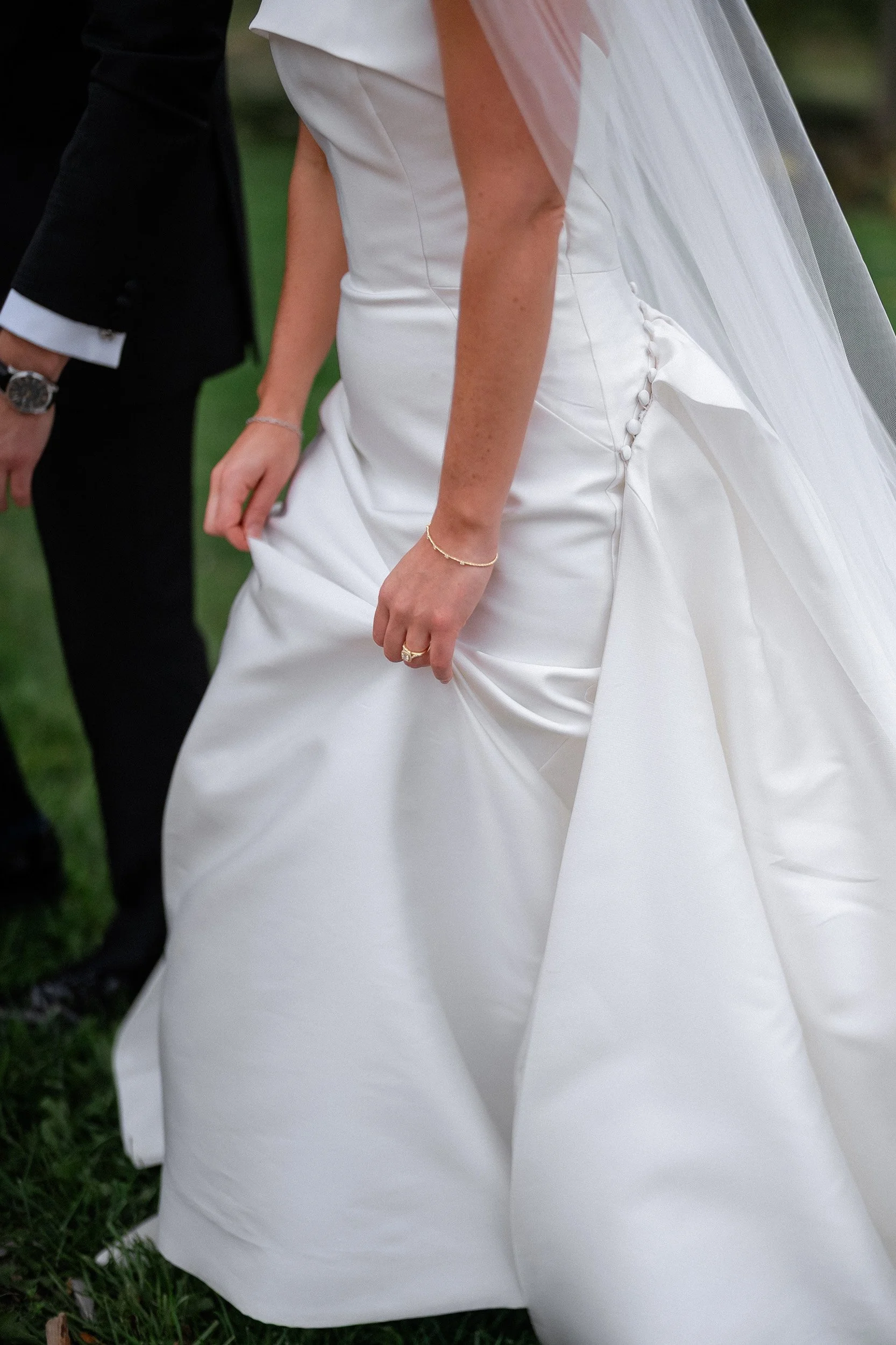 Close-up of a bride in a white wedding gown holding the skirt, with a groom in a black suit standing beside her.