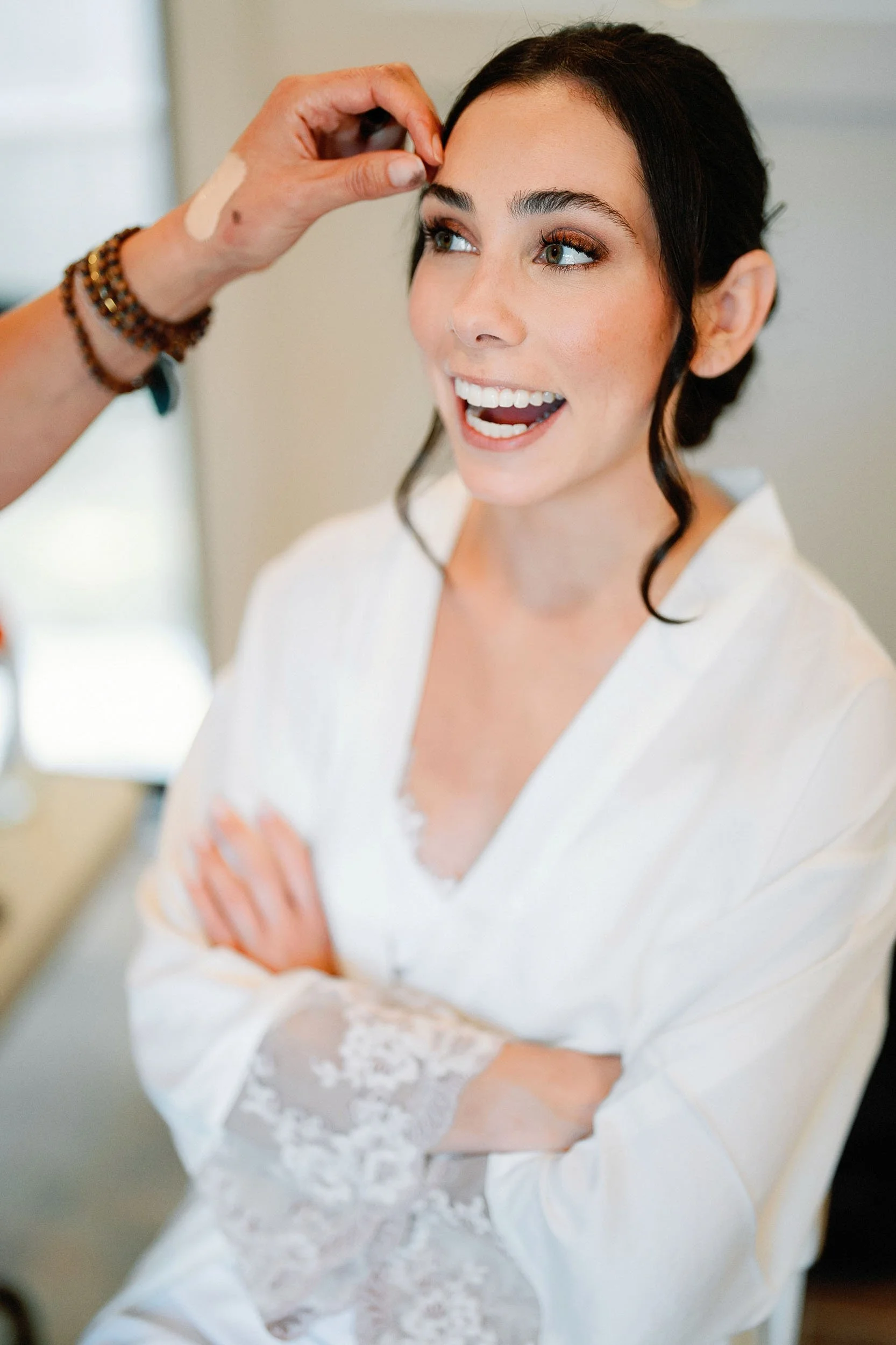 Woman with dark hair smiling as makeup artist applies makeup, wearing a white robe with lace sleeves.