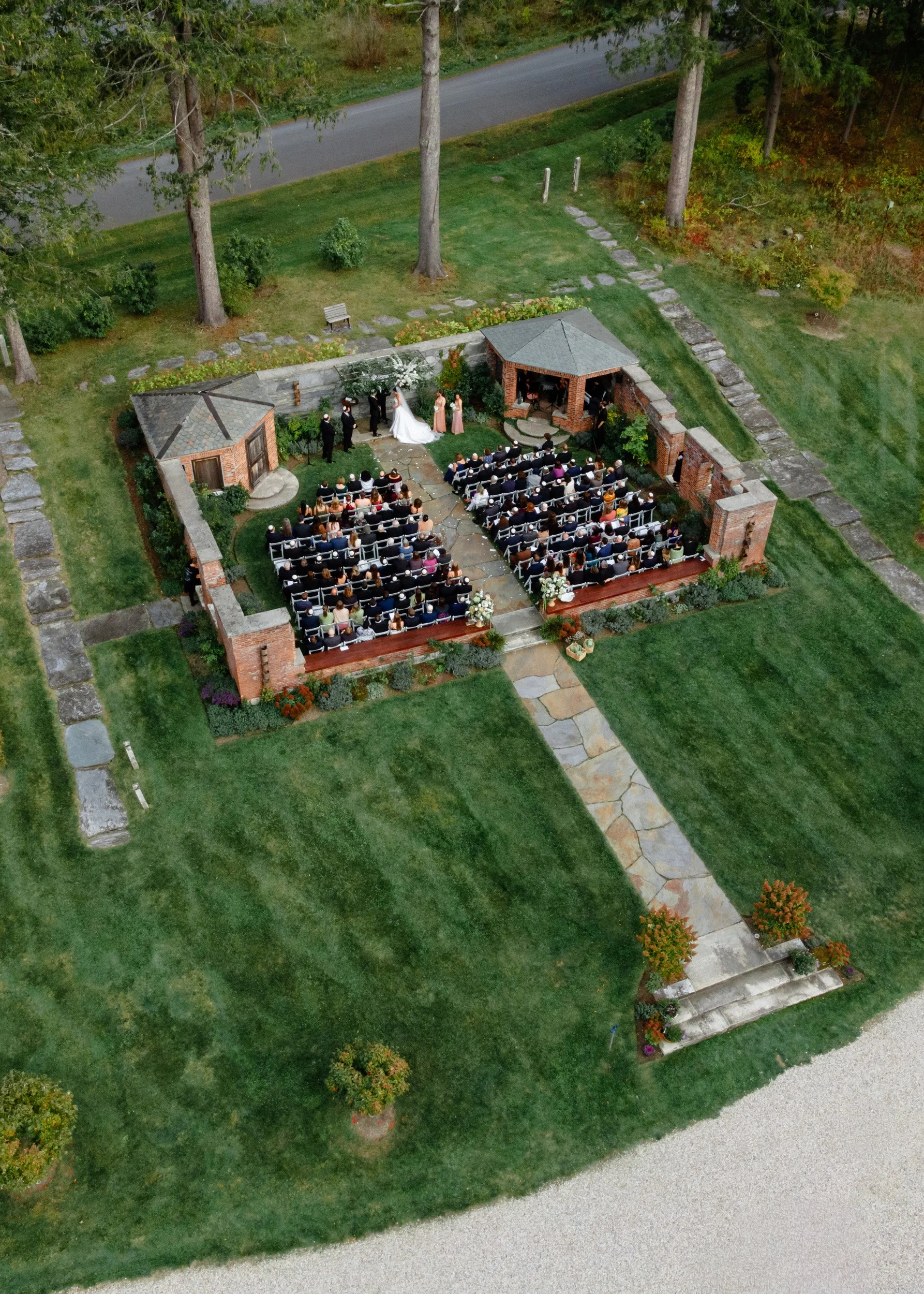 Aerial view of a small outdoor wedding ceremony in a lush green garden, surrounded by trees, with guests seated on white chairs, a bride and groom at the altar, and a small brick pavilion.