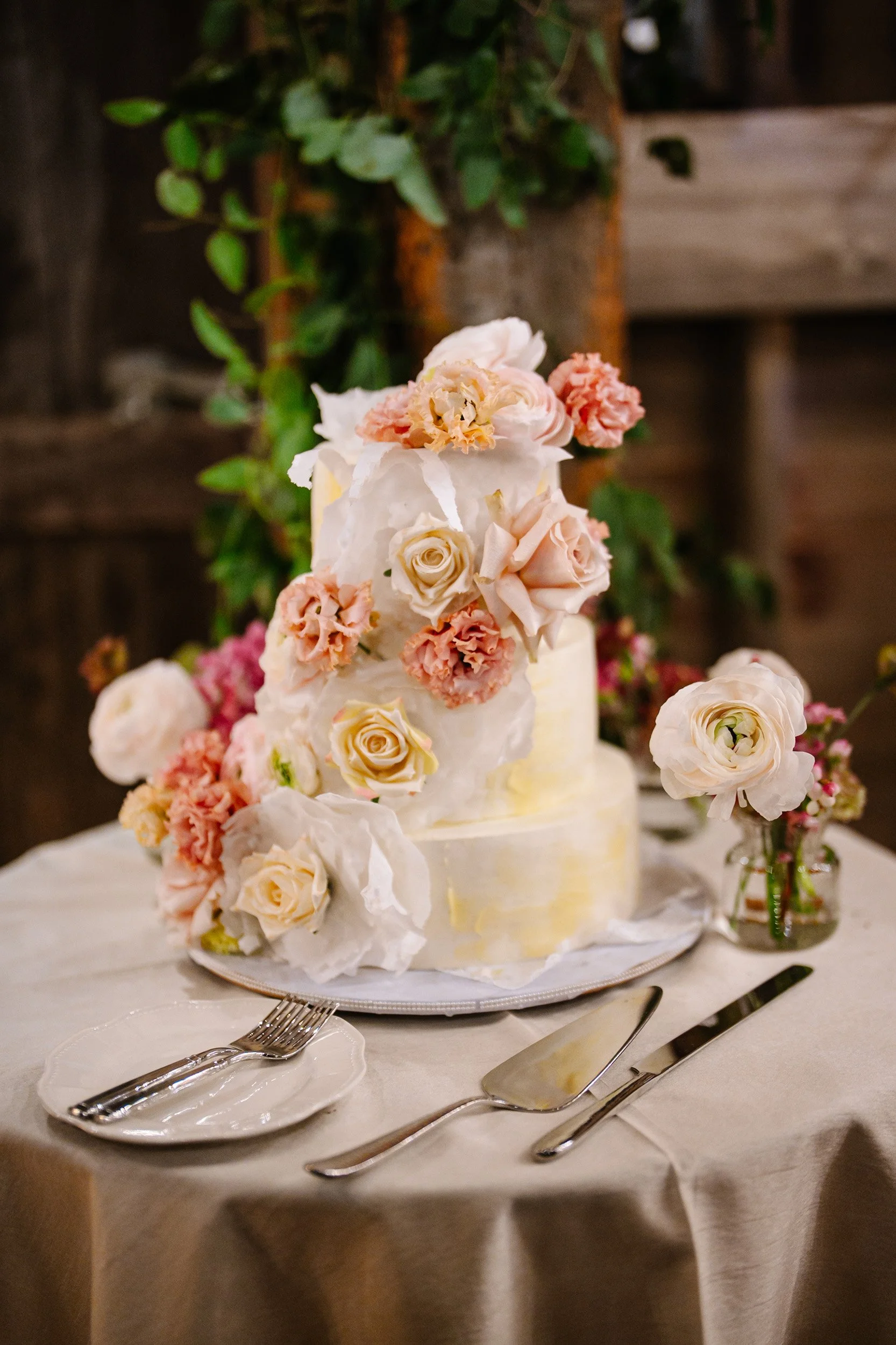 A wedding cake decorated with white and peach roses and pink blossoms, set on a round table with cream-colored tablecloth, silverware, and small vases with flowers, against a wooden background.
