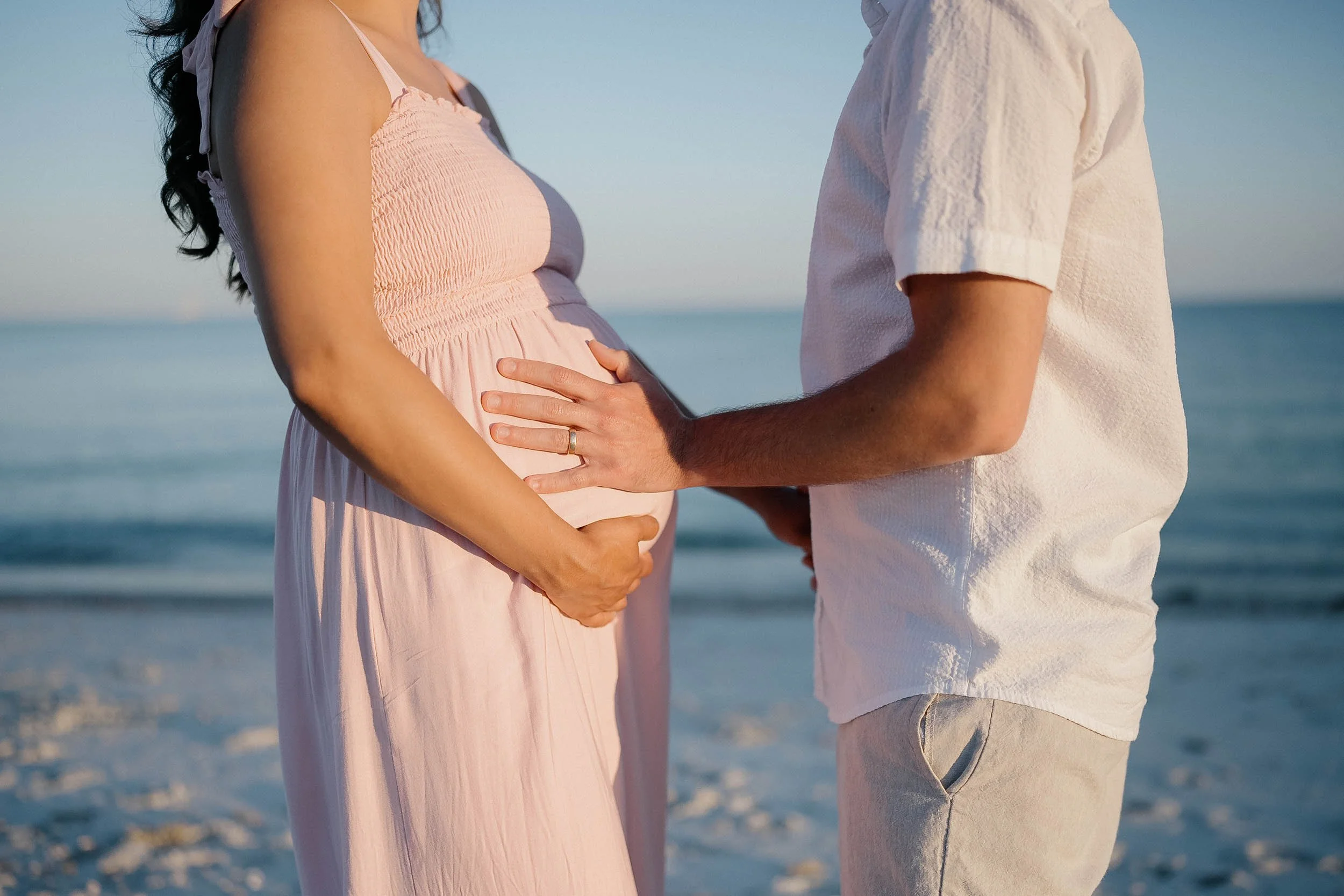 A pregnant woman in a pink dress holding her belly, standing beside a man at the beach, who is touching her belly with one hand.