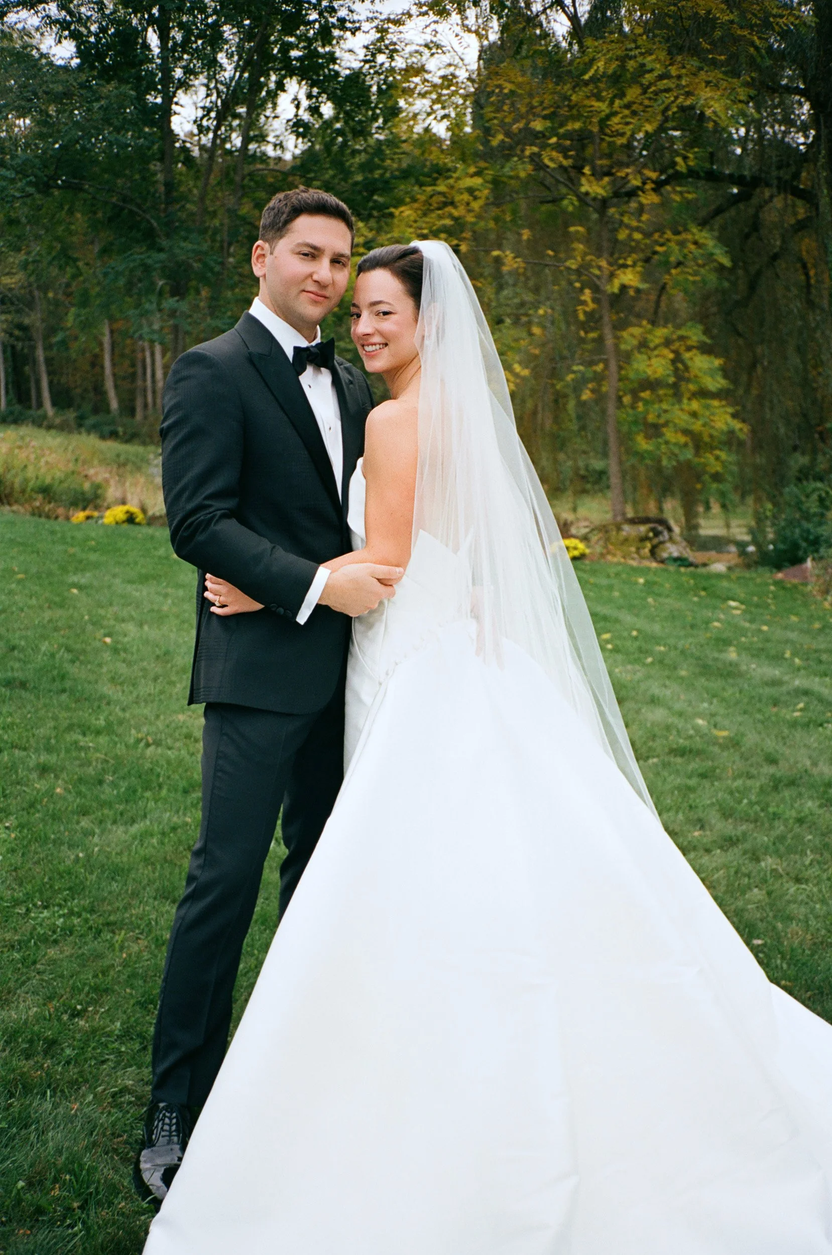 A bride and groom standing together outdoors during daytime, with trees and greenery in the background. The bride wears a white wedding dress with a veil, and the groom wears a black tuxedo with a bowtie.