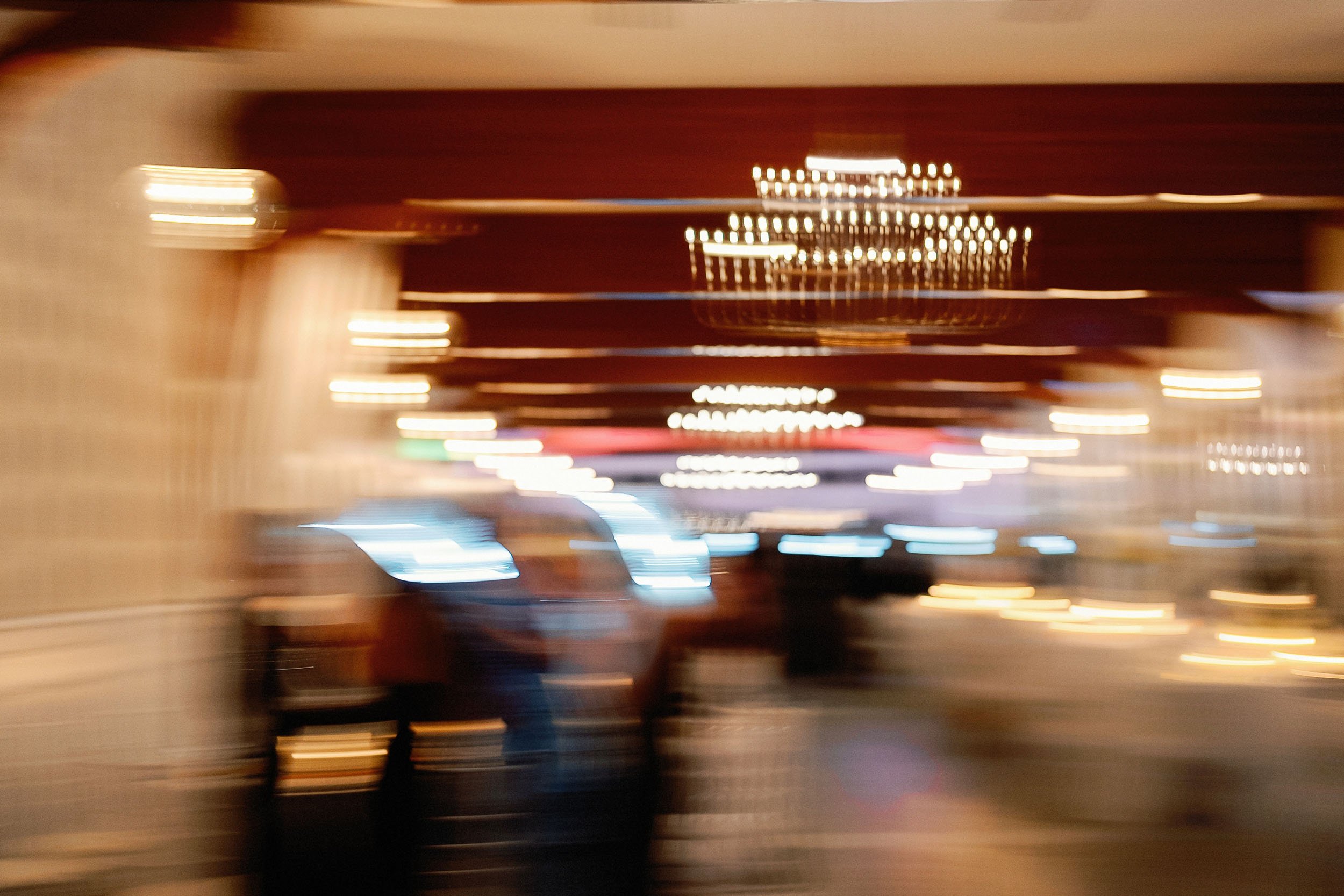 Blurred image of a casino or gaming area with chandelier and multicolored lights, showing people walking and standing.