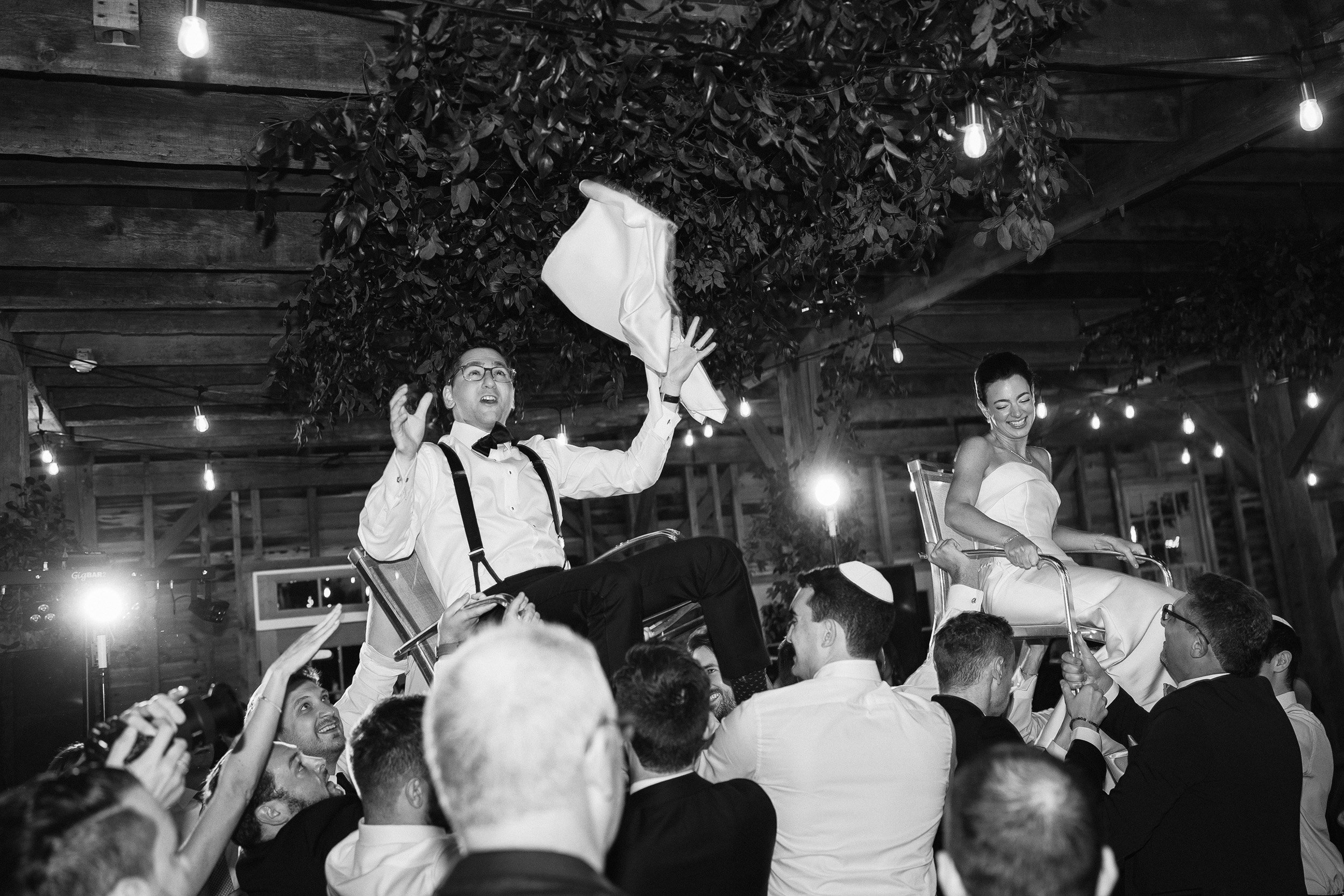 People celebrating at a wedding, with the bride and groom on chairs being lifted by guests in a rustic venue decorated with string lights and greenery, in black and white.