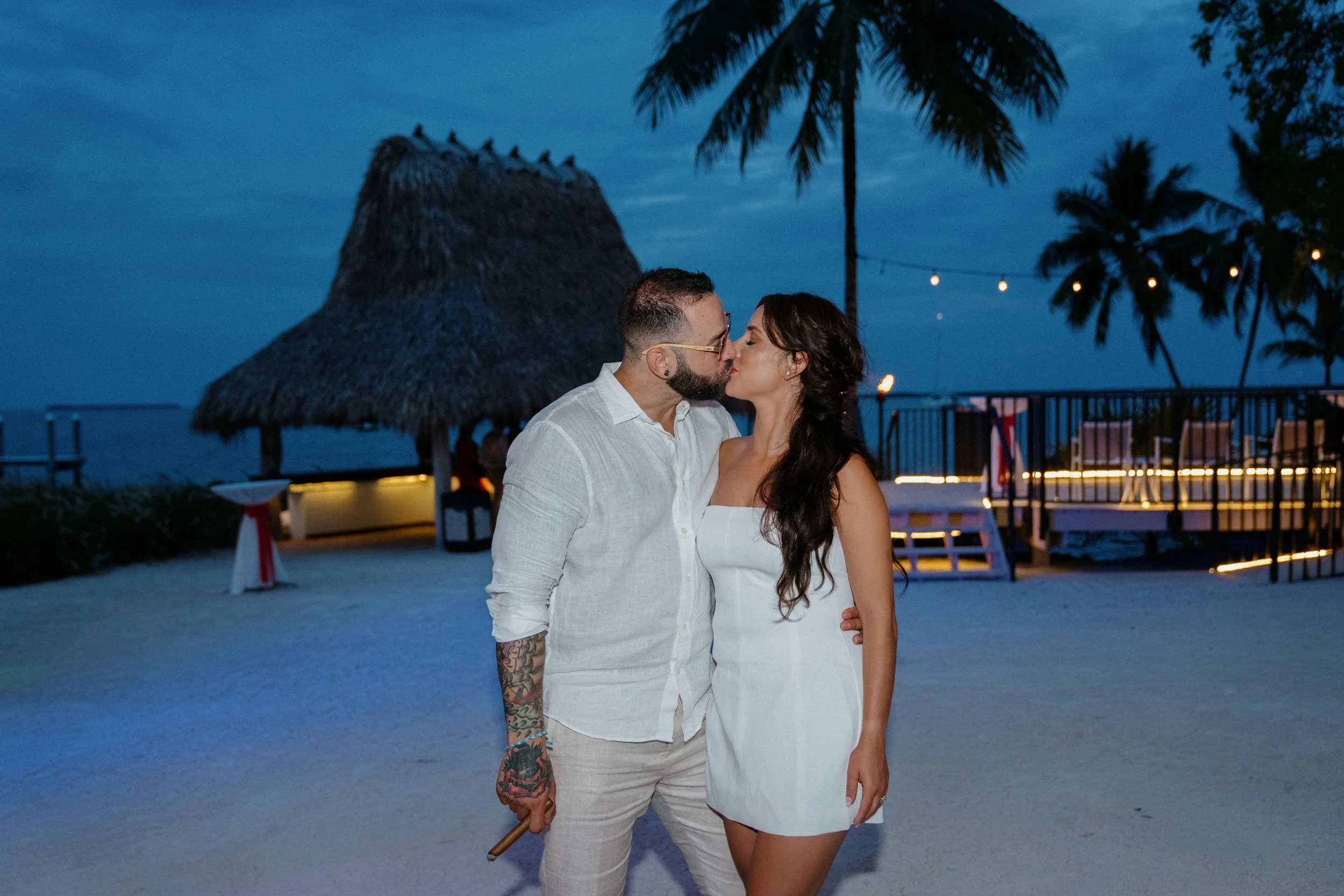 A couple sharing a kiss on a beach at dusk, with palm trees, a thatched hut, and string lights in the background.