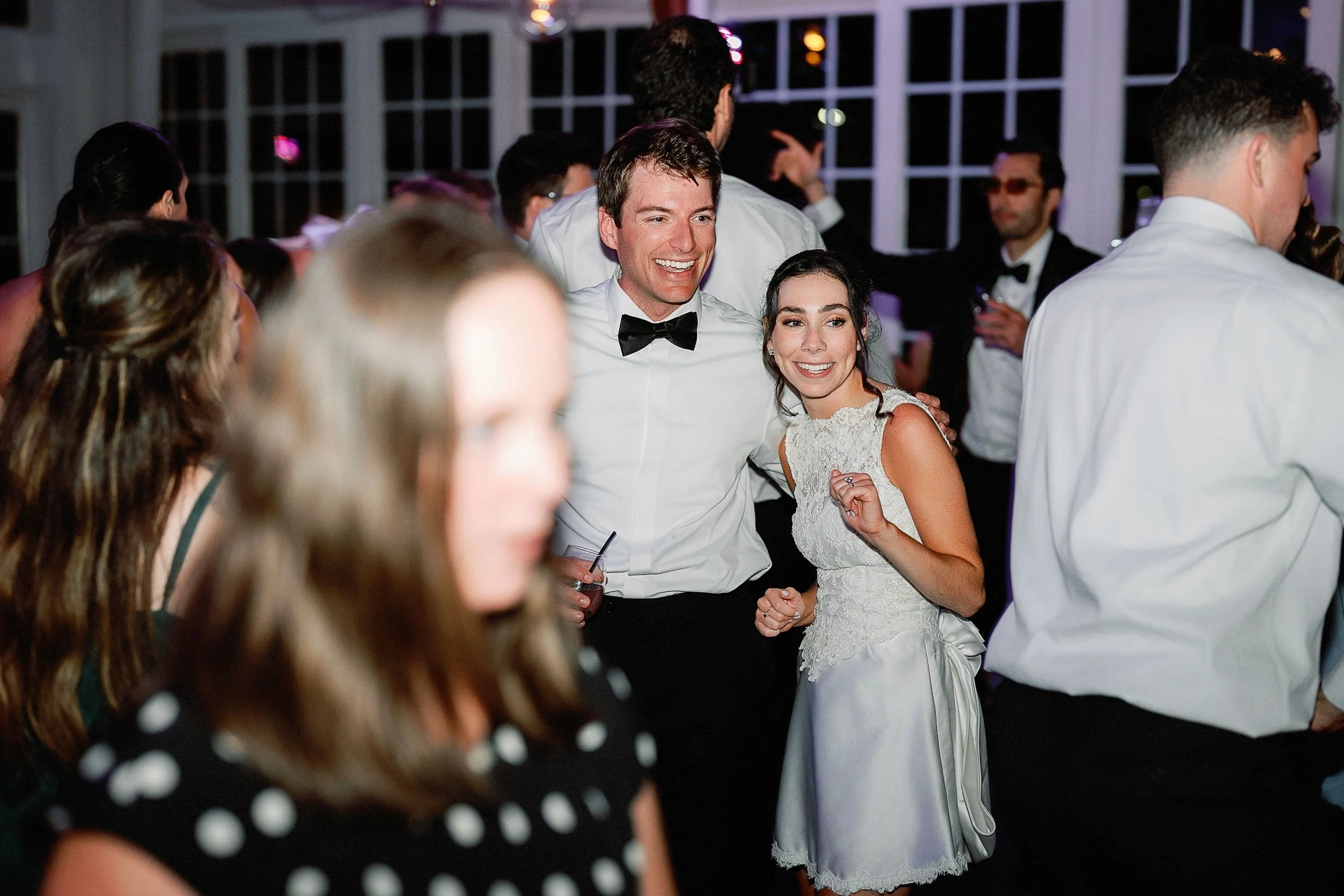A group of people dancing at a wedding reception, with a man in a tuxedo and a woman in a white dress smiling and holding hands.