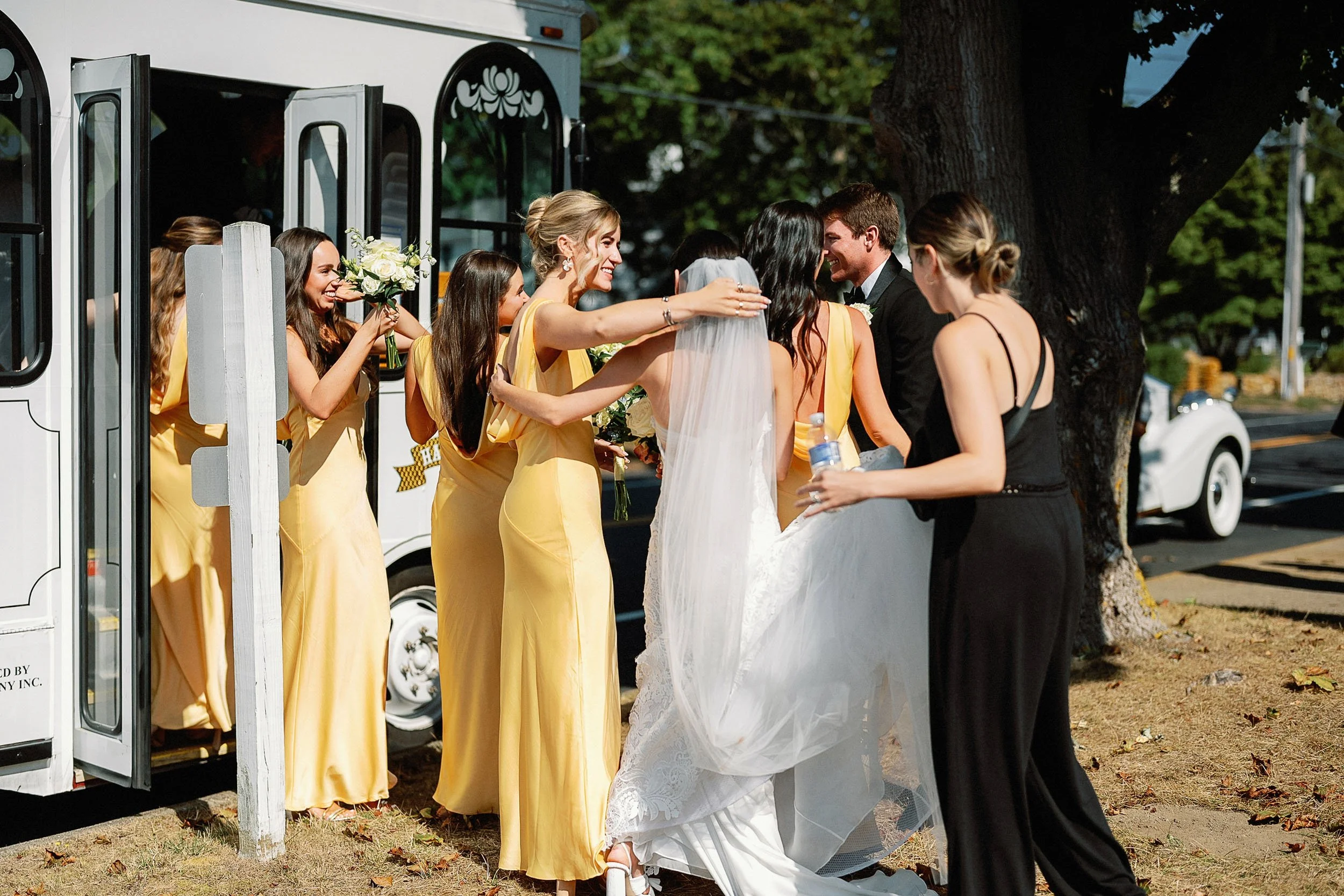 A bride in a white wedding dress and veil hugging a bridesmaid in a yellow dress as she arrives at her wedding. Several bridesmaids in yellow dresses and a woman in black dress are standing near a white vehicle with an open door, outdoors near a larg
