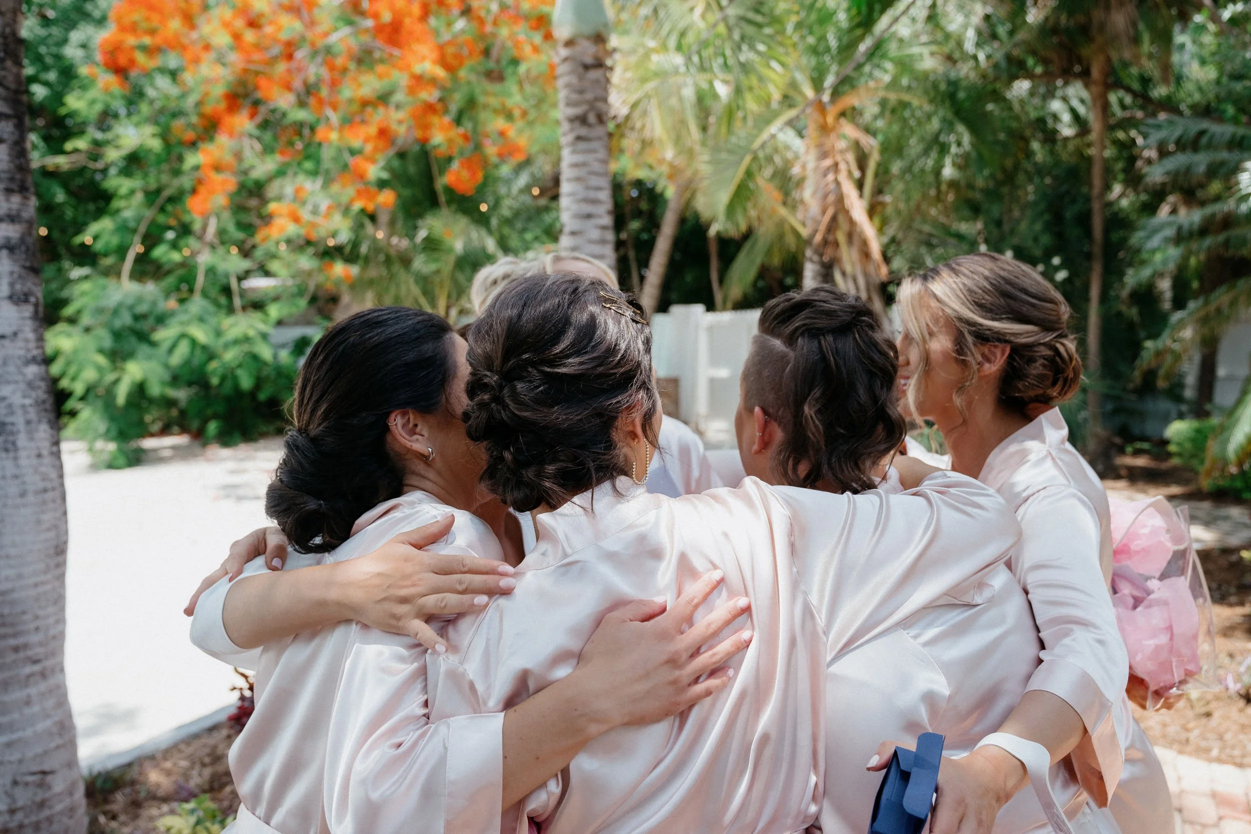 Group of women hugging outdoors among trees and tropical plants.