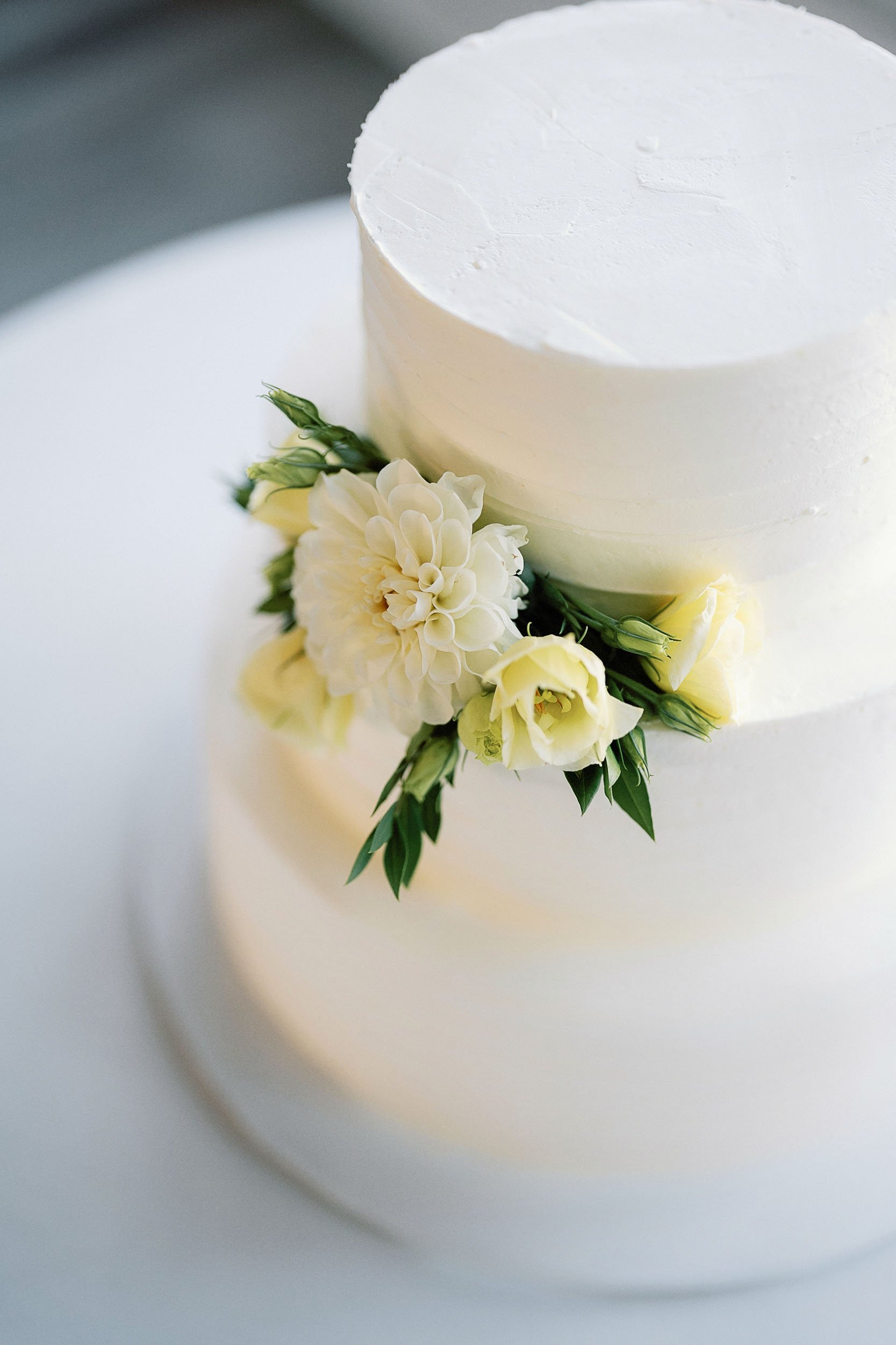 A three-tiered white wedding cake decorated with white flowers and green leaves.