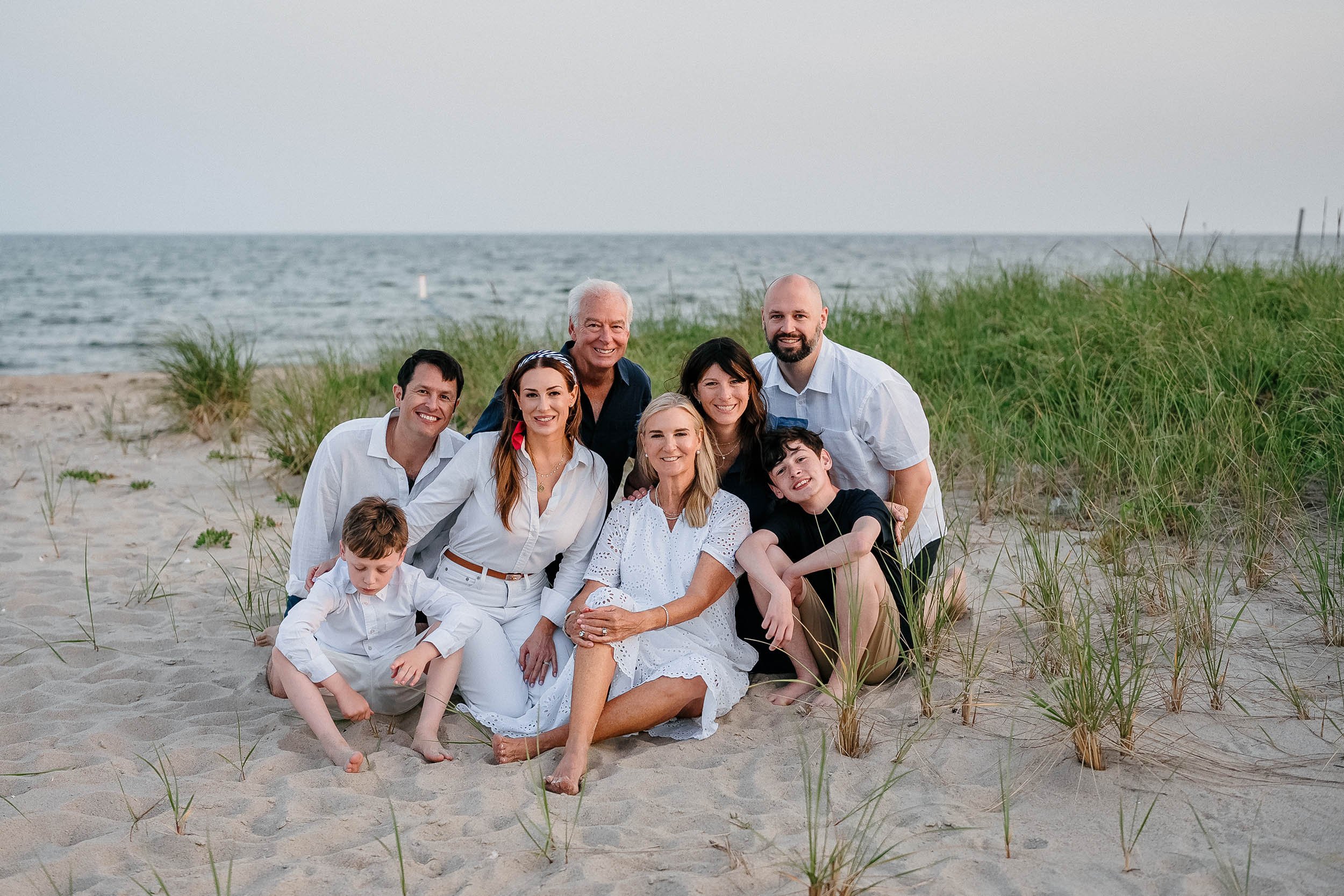 A large family of nine people on a sandy beach with ocean and grassy dunes in the background, smiling and posing for a group photo.
