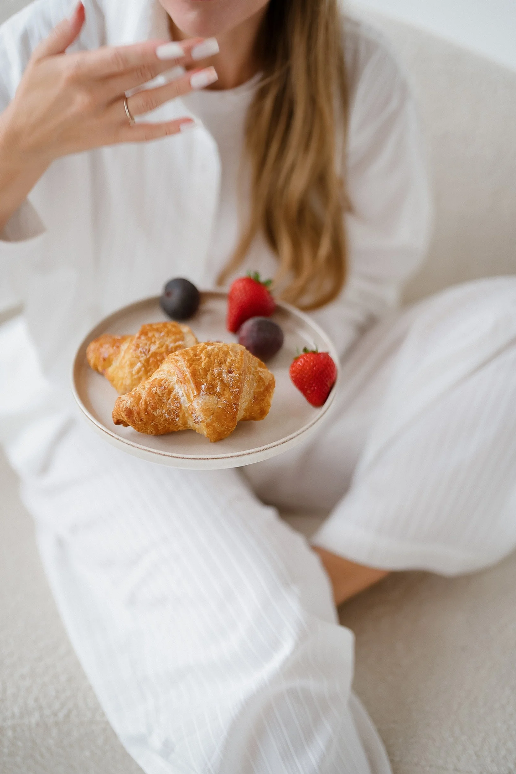 A woman in white pajamas is sitting, holding a small plate with two croissants and fresh strawberries and blueberries, while sipping a beverage.