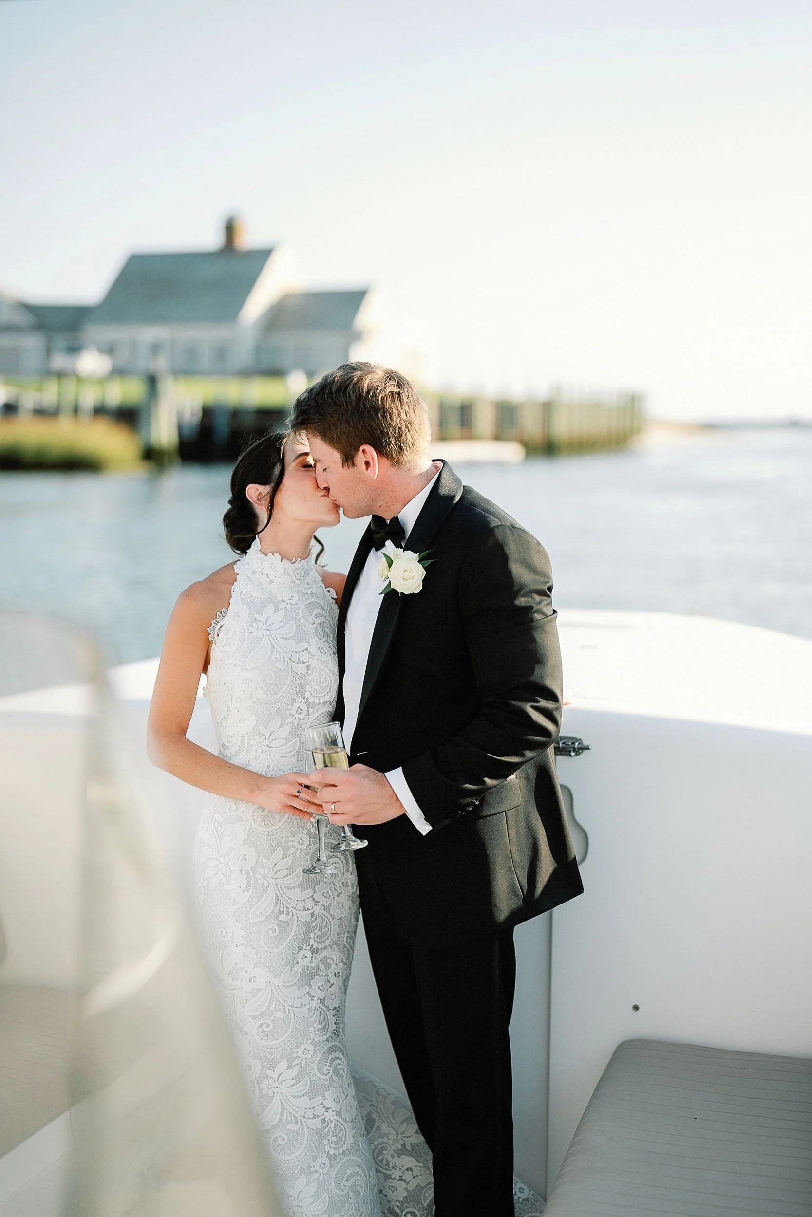 A bride and groom sharing a kiss on a boat with water and houses in the background during sunset, the bride holding a glass of champagne.