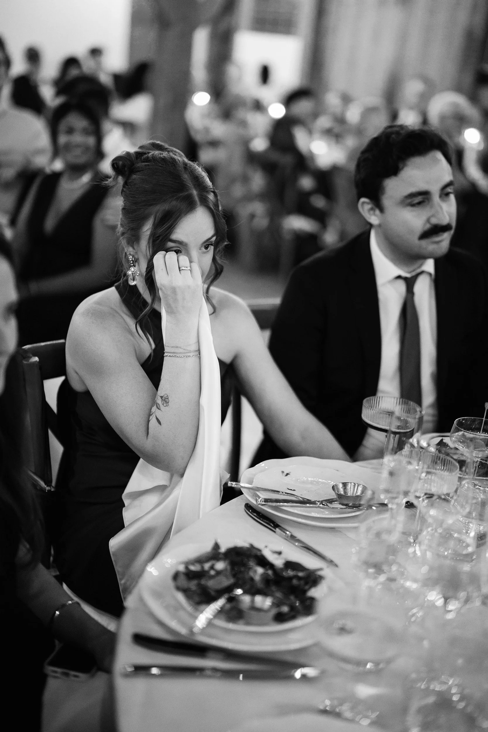 A woman with wavy hair, earrings, and a tattoo on her arm, wiping her eye, sitting at a table with a man dressed in a suit, in a formal setting with other guests in the background.
