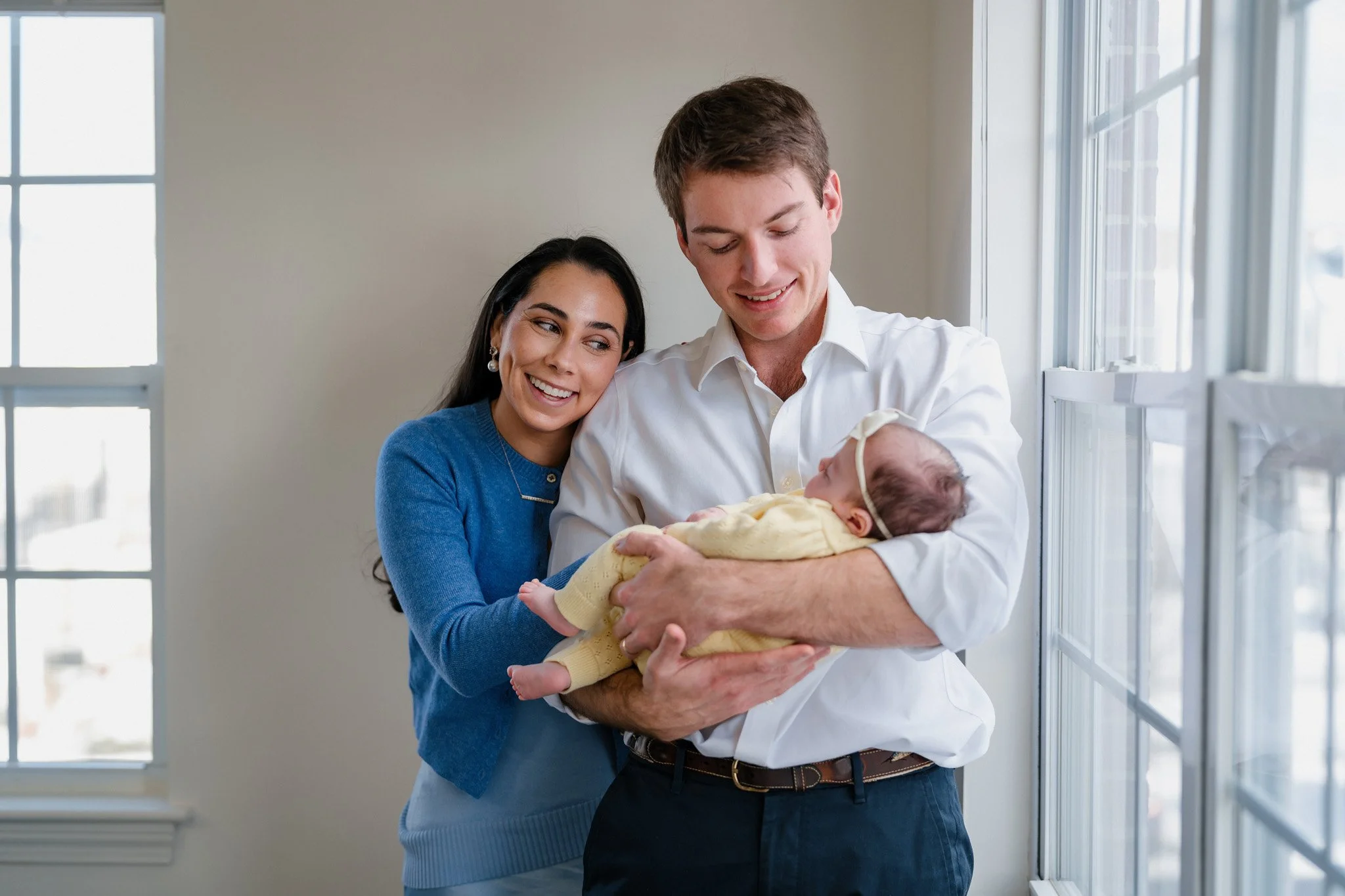 A happy couple holding a baby near a window in a well-lit room.