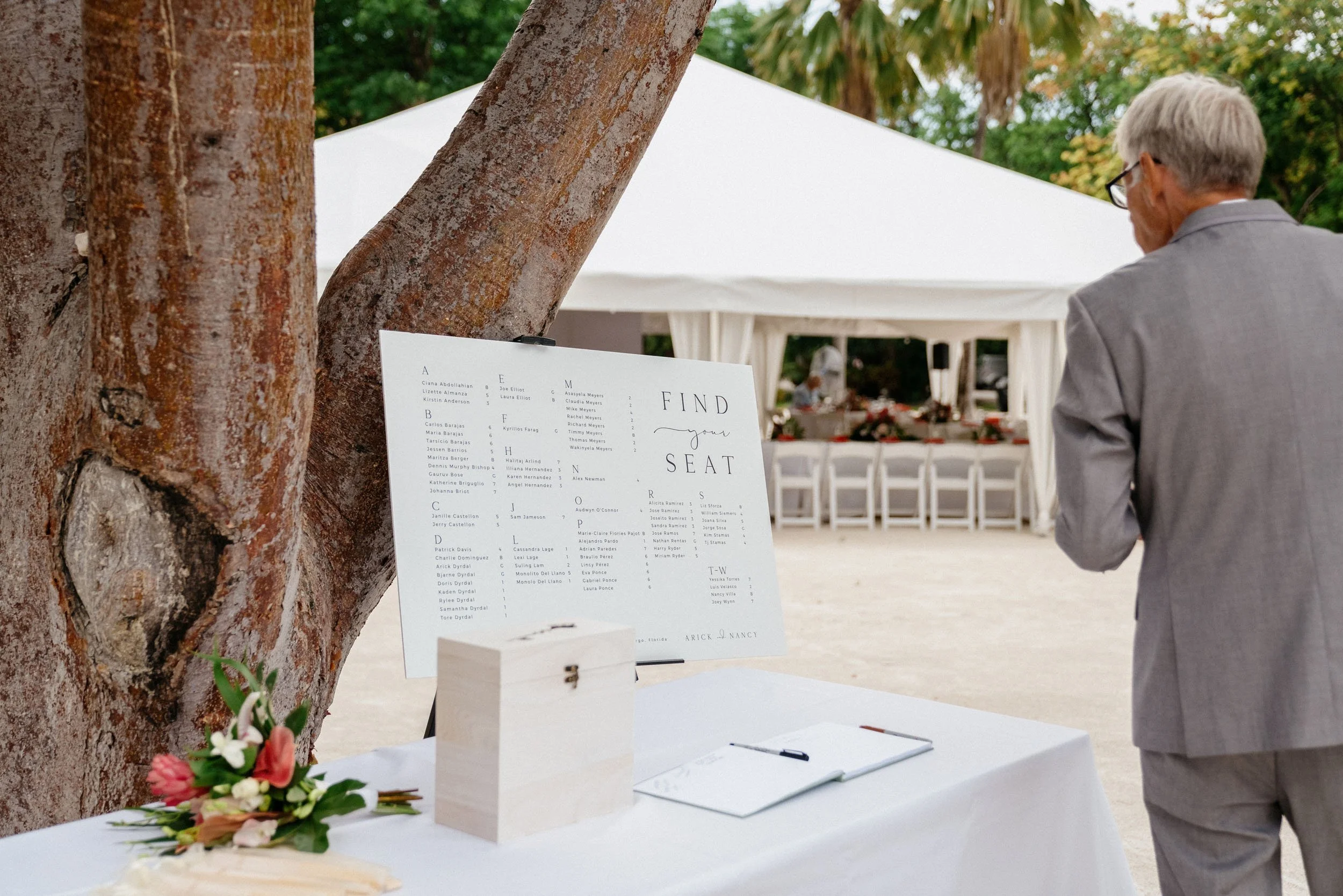 A man in a gray suit standing outside at a wedding reception, looking at a seating chart display on an easel at a table decorated with a bouquet of flowers and a wooden box.