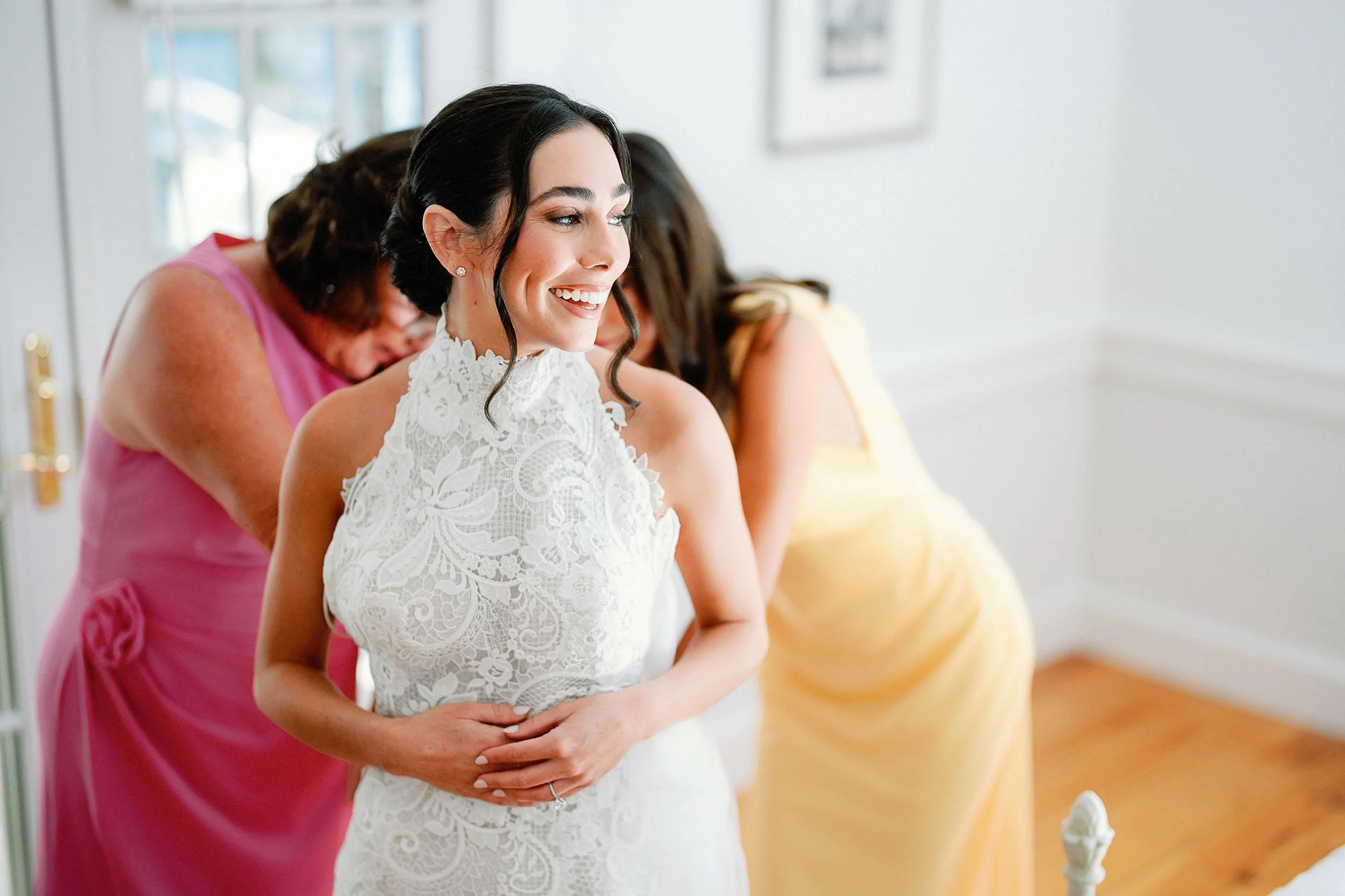 A smiling bride in a white lace wedding dress, two women in yellow and pink dresses helping her get ready indoors with framed pictures on white walls in the background.