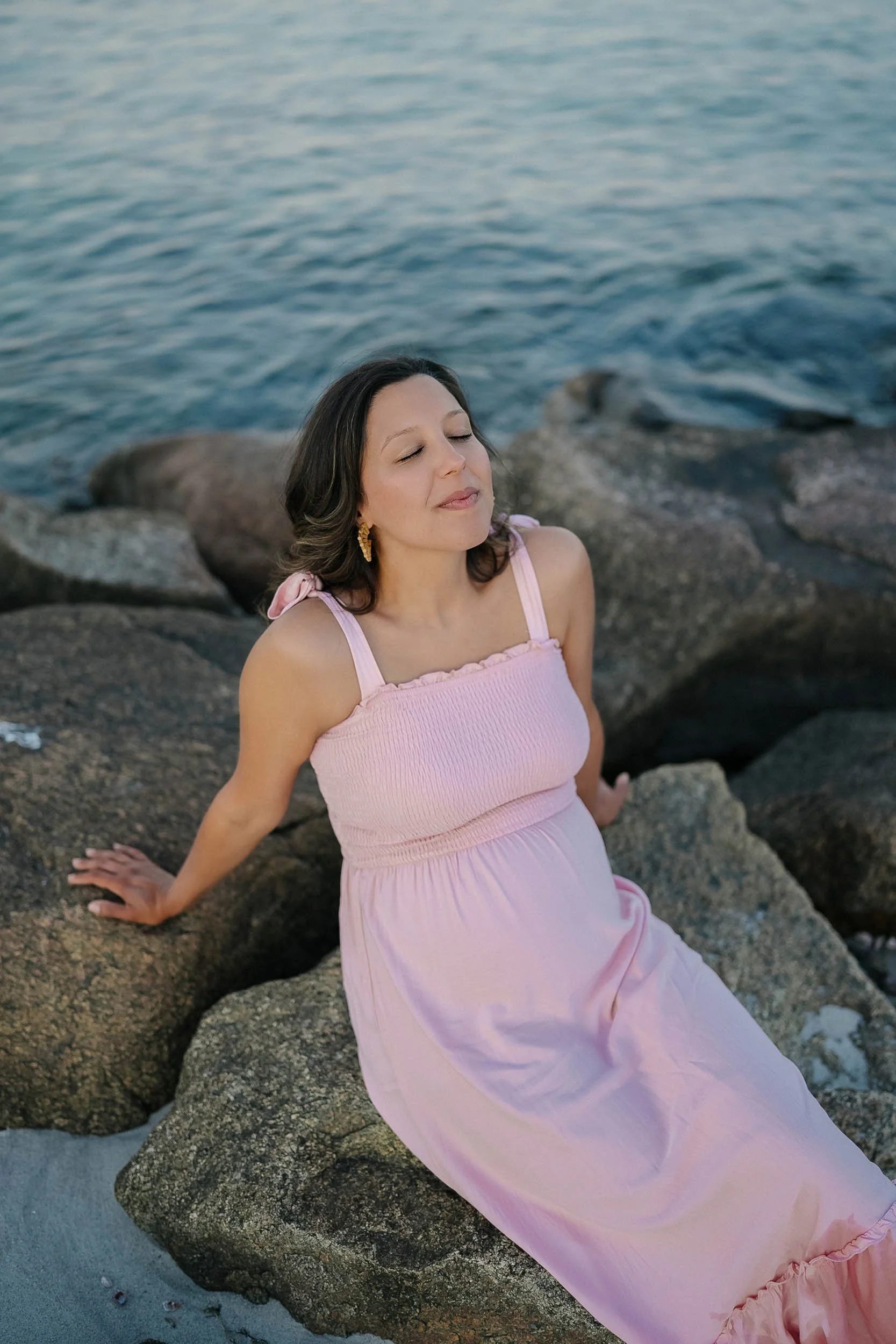 A woman in a pink dress sitting on rocks by the water with her eyes closed and a peaceful expression.