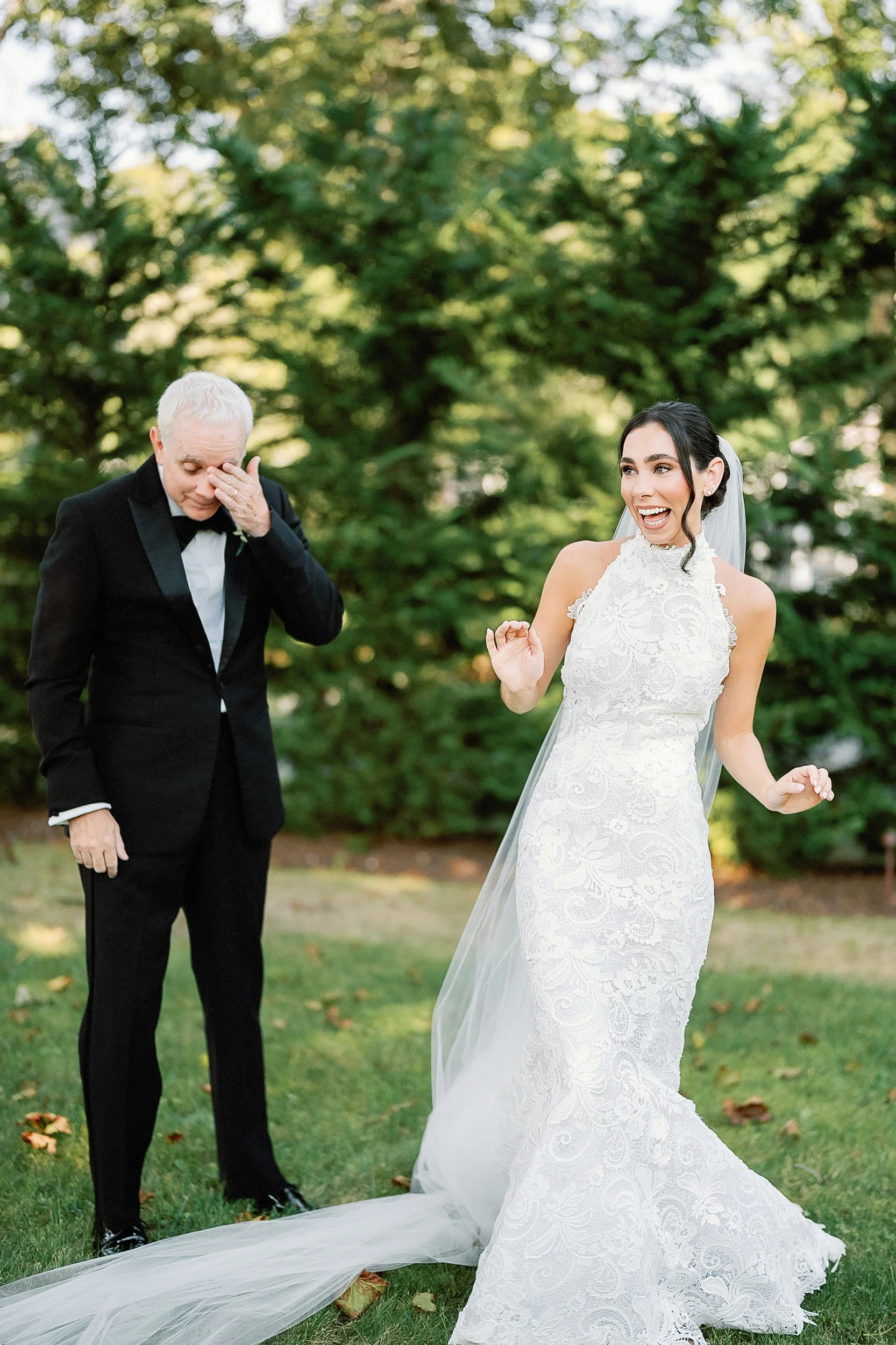 A bride in a white lace wedding gown and veil, smiling and gesturing happily, standing outdoors on grass with a groom in a black tuxedo wiping away tears, surrounded by green trees.