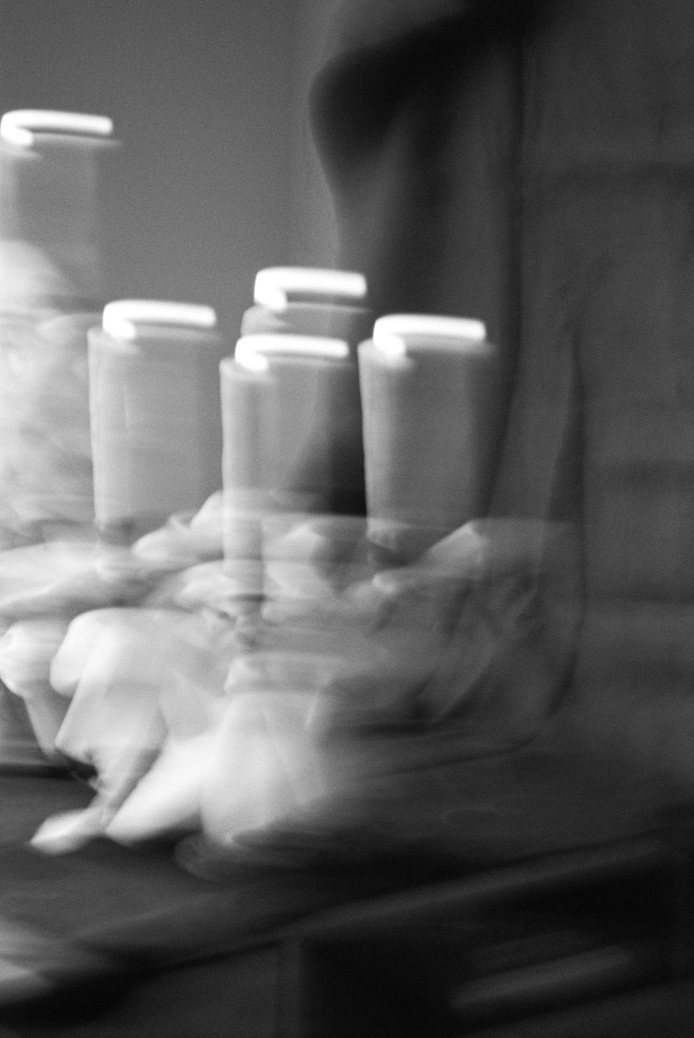 Blurry black and white image of at least five cans or bottles with white caps on a surface, with a person’s hand reaching towards them.