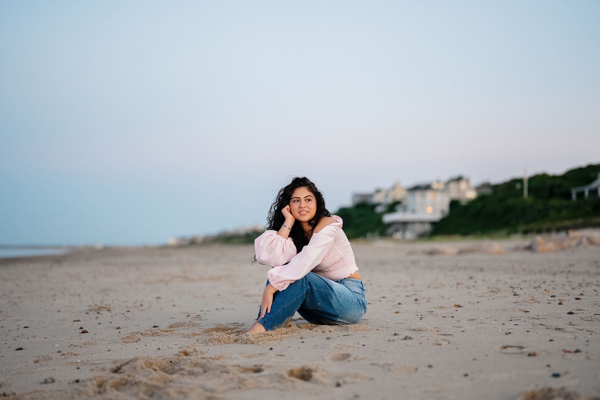 Young woman sitting on the sandy beach with houses and greenery in the background during early evening or late afternoon.