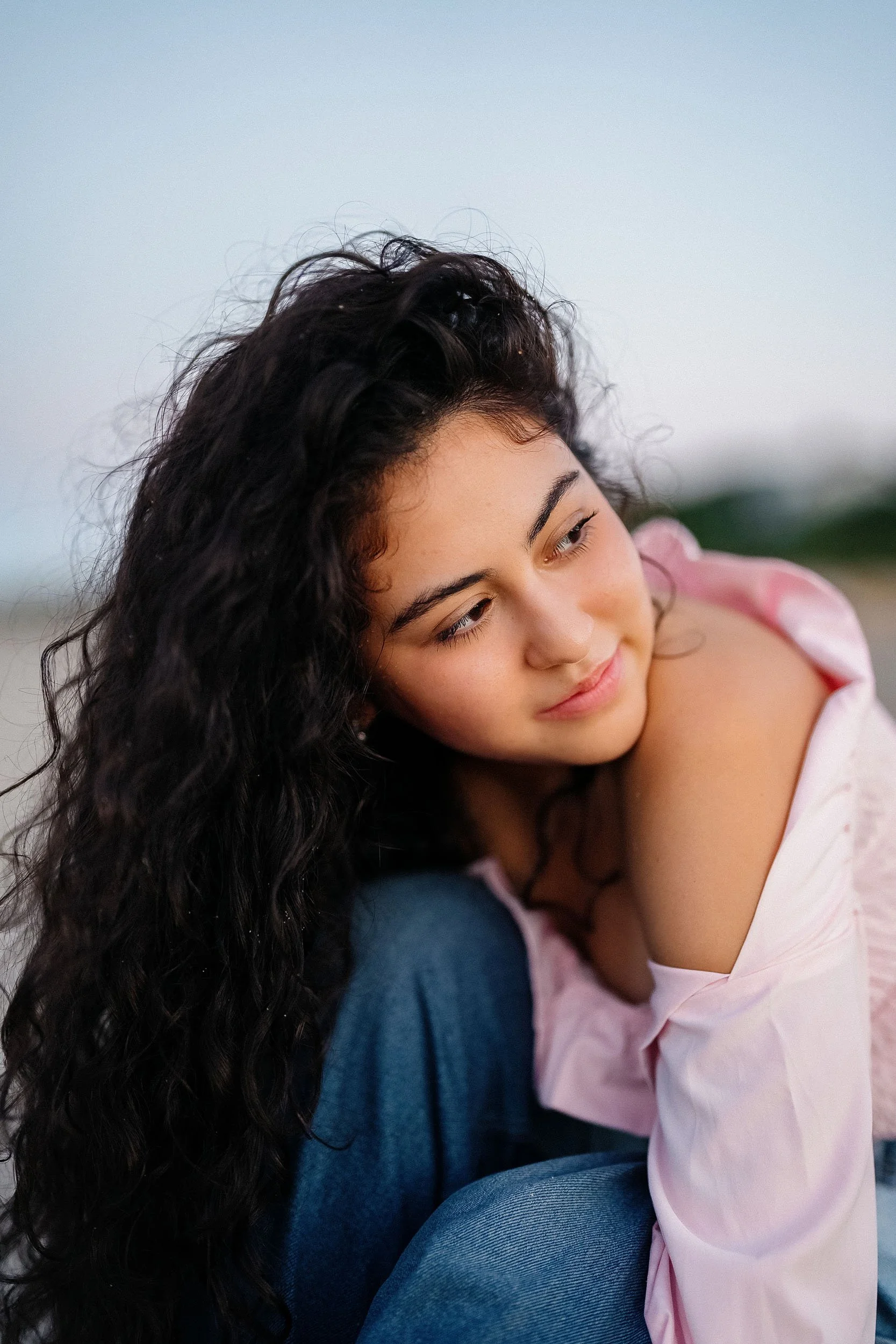 A young woman with long curly dark hair and light skin, sitting outdoors, looking thoughtfully downward, wearing a pink off-shoulder top and blue jeans.