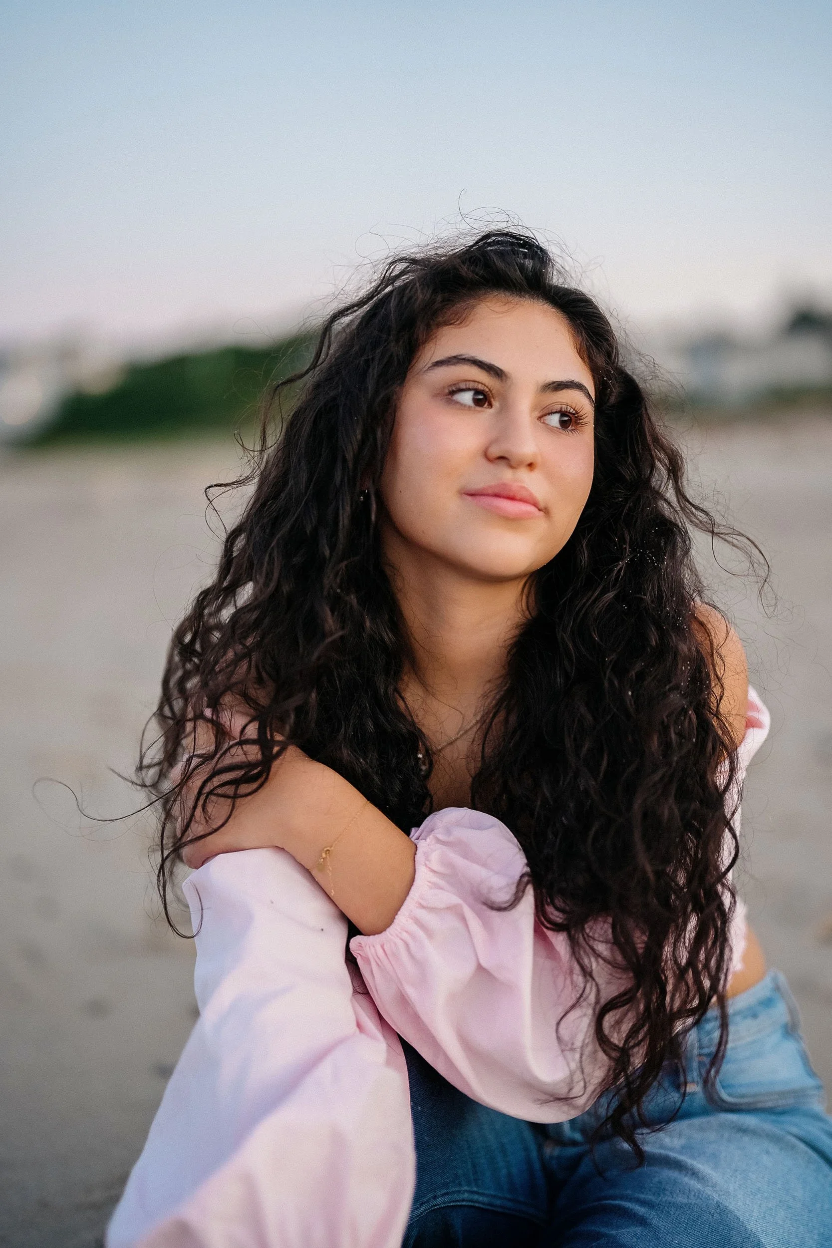 A young woman with long, curly dark hair sitting on the beach, wearing a light pink jacket and blue jeans, gazing thoughtfully into the distance during sunset.
