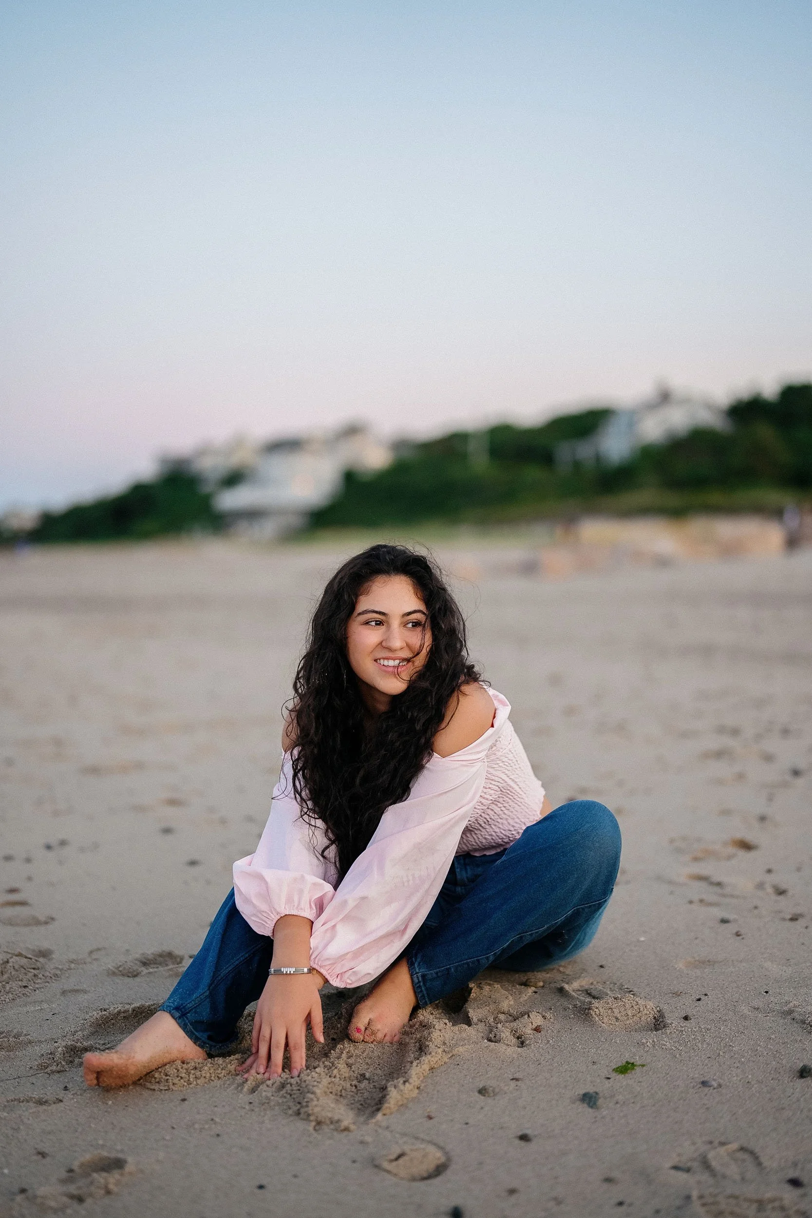 A young woman with long curly dark hair sitting on the sandy beach with her legs crossed, smiling and looking to the side, wearing a pink off-shoulder top and blue jeans, with houses and greenery in the background.