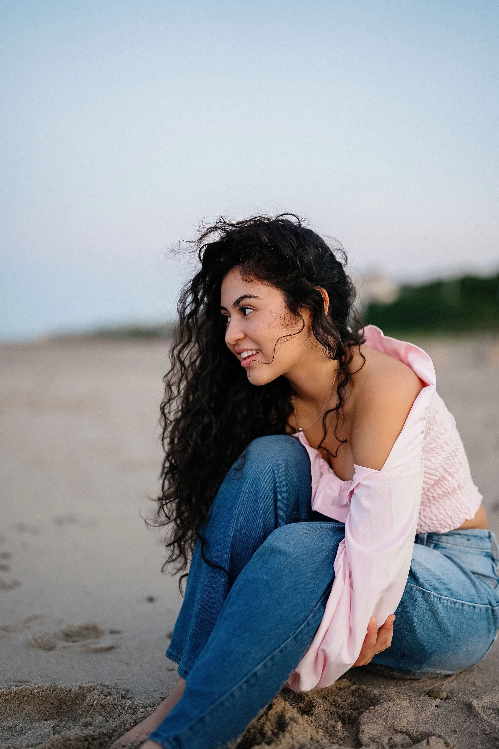 A young woman with long, curly dark hair sitting on a sandy beach during sunset, wearing a light pink off-shoulder top and blue jeans, smiling and looking to her left.