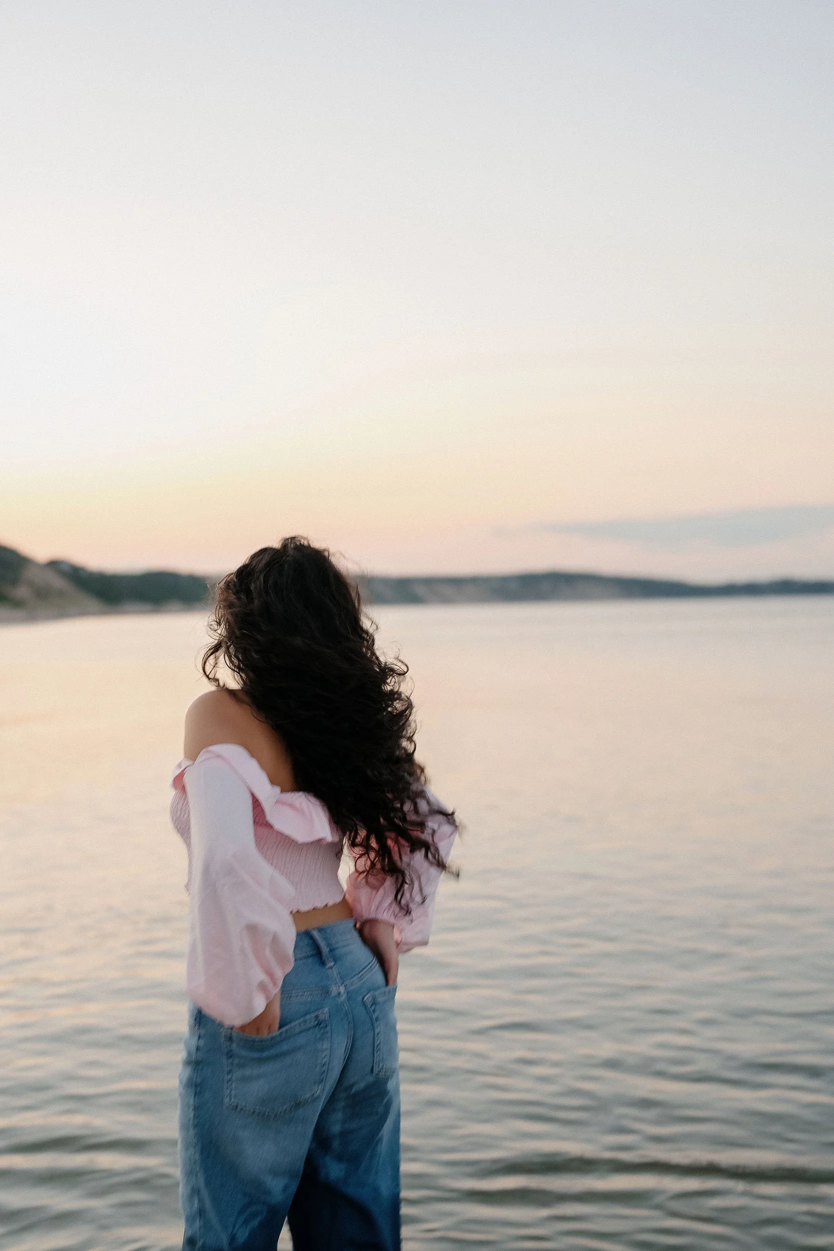 A woman with long, curly black hair standing by water during sunset, wearing a pink off-shoulder top and blue jeans.