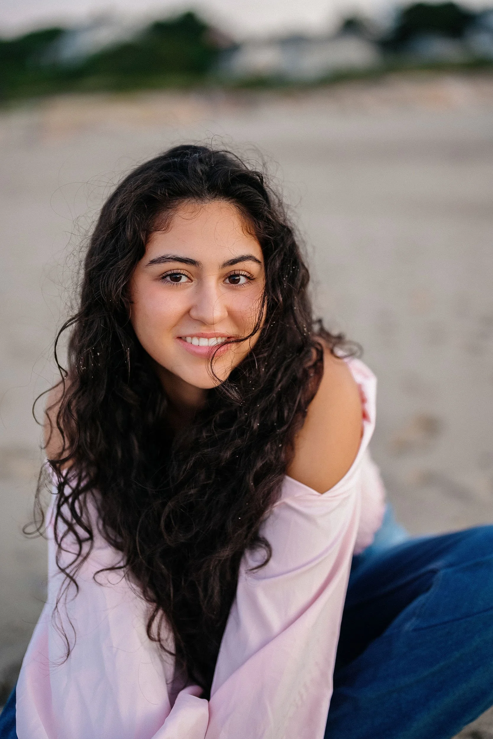A young woman with long, dark, curly hair smiling at the camera, sitting on the beach in casual clothing.