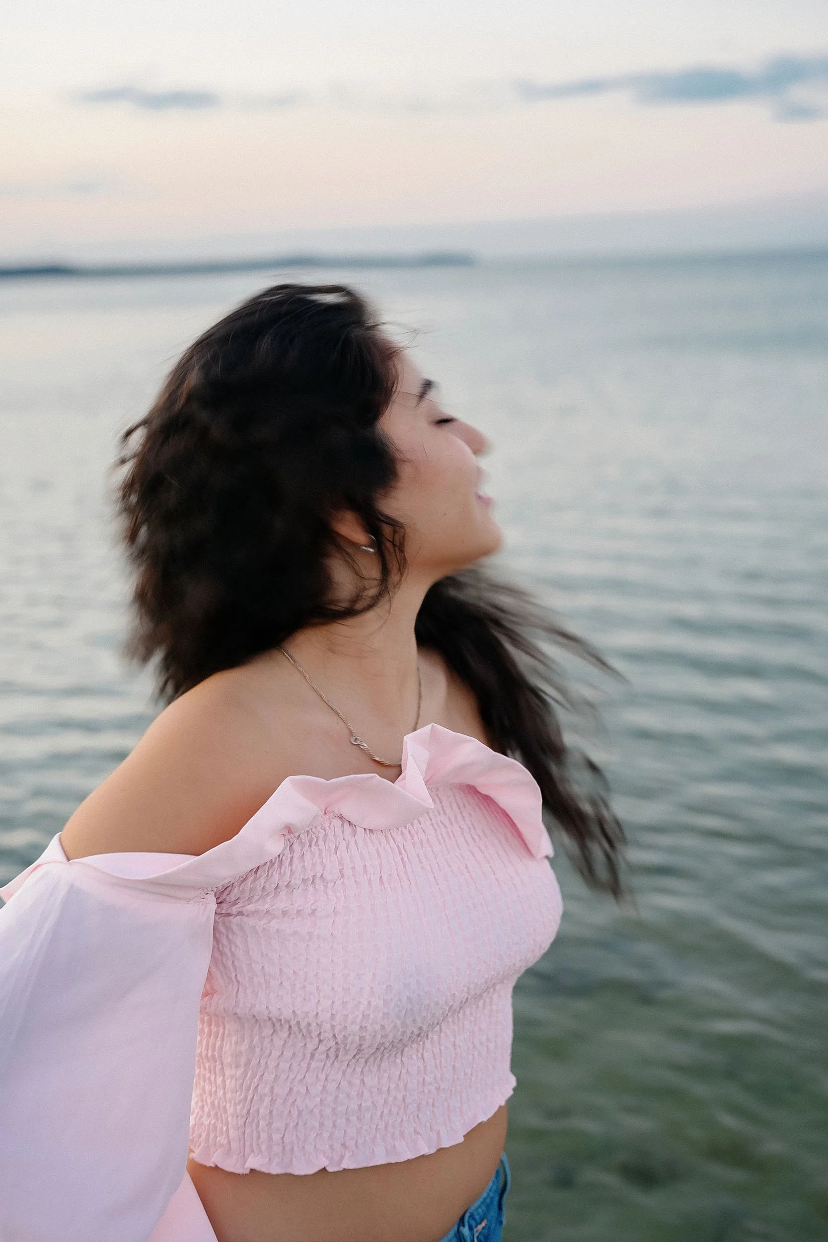 A woman with long dark hair is standing by the water, facing sideways with eyes closed, wearing a pink off-the-shoulder top and a silver necklace, during sunset or sunrise.
