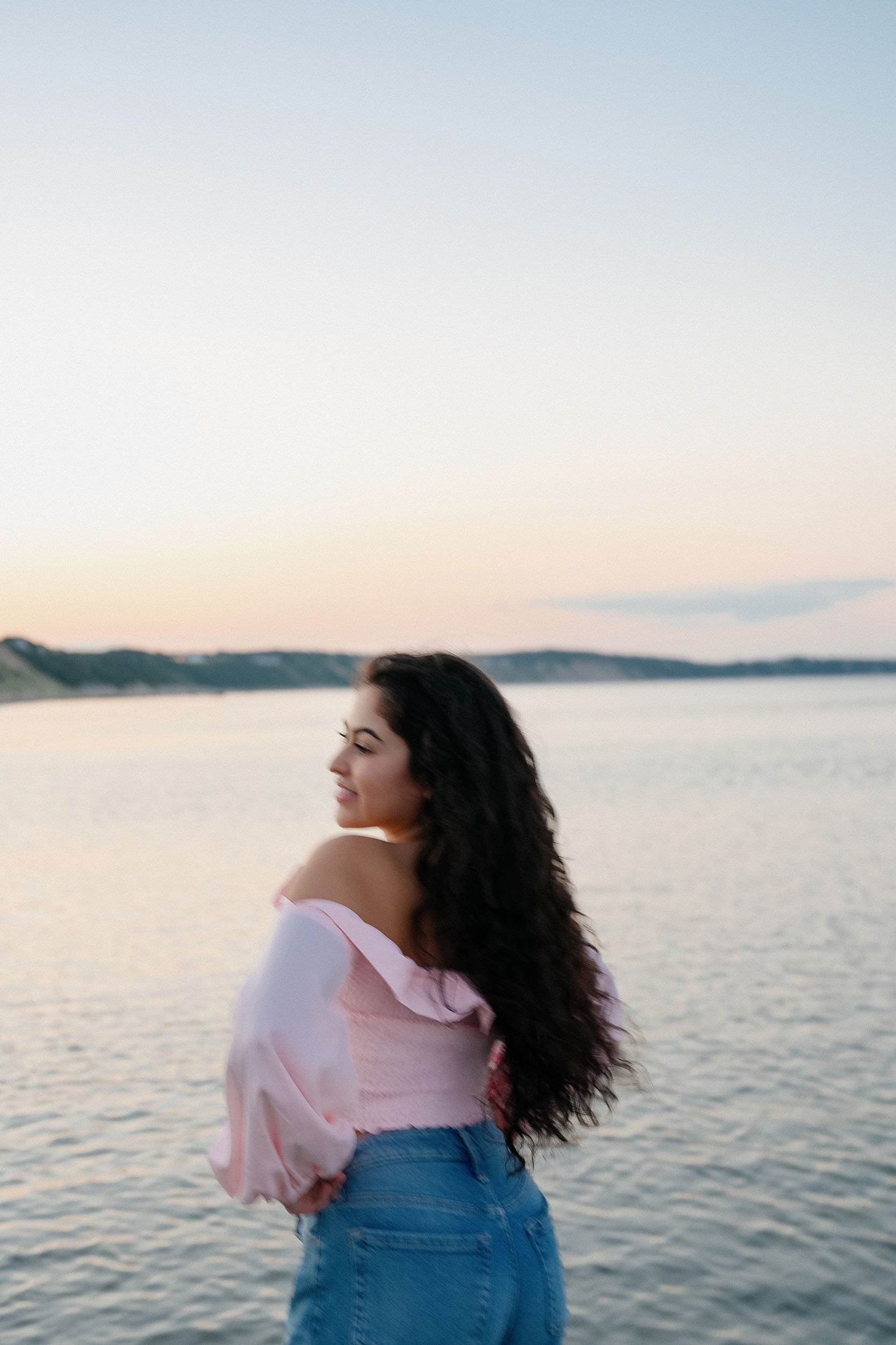 A woman with long, curly dark hair standing by a body of water during sunset, wearing an off-the-shoulder pink top and blue jeans, smiling with her eyes closed.