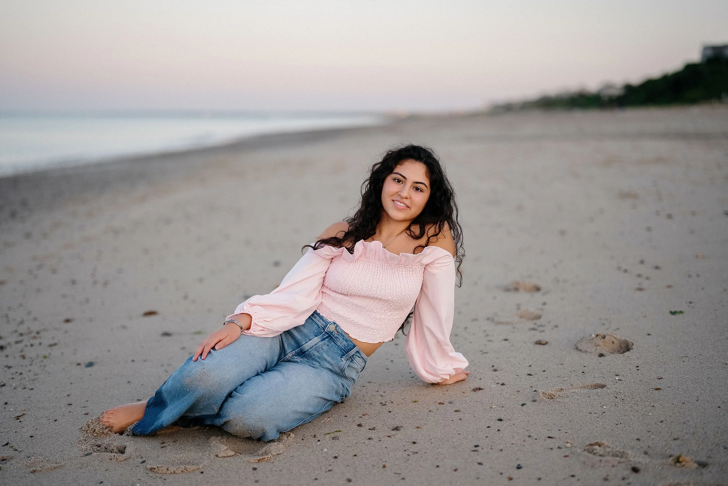 A woman with long dark curly hair sitting on a sandy beach near the ocean during dusk, wearing a pink off-shoulder top and blue jeans.