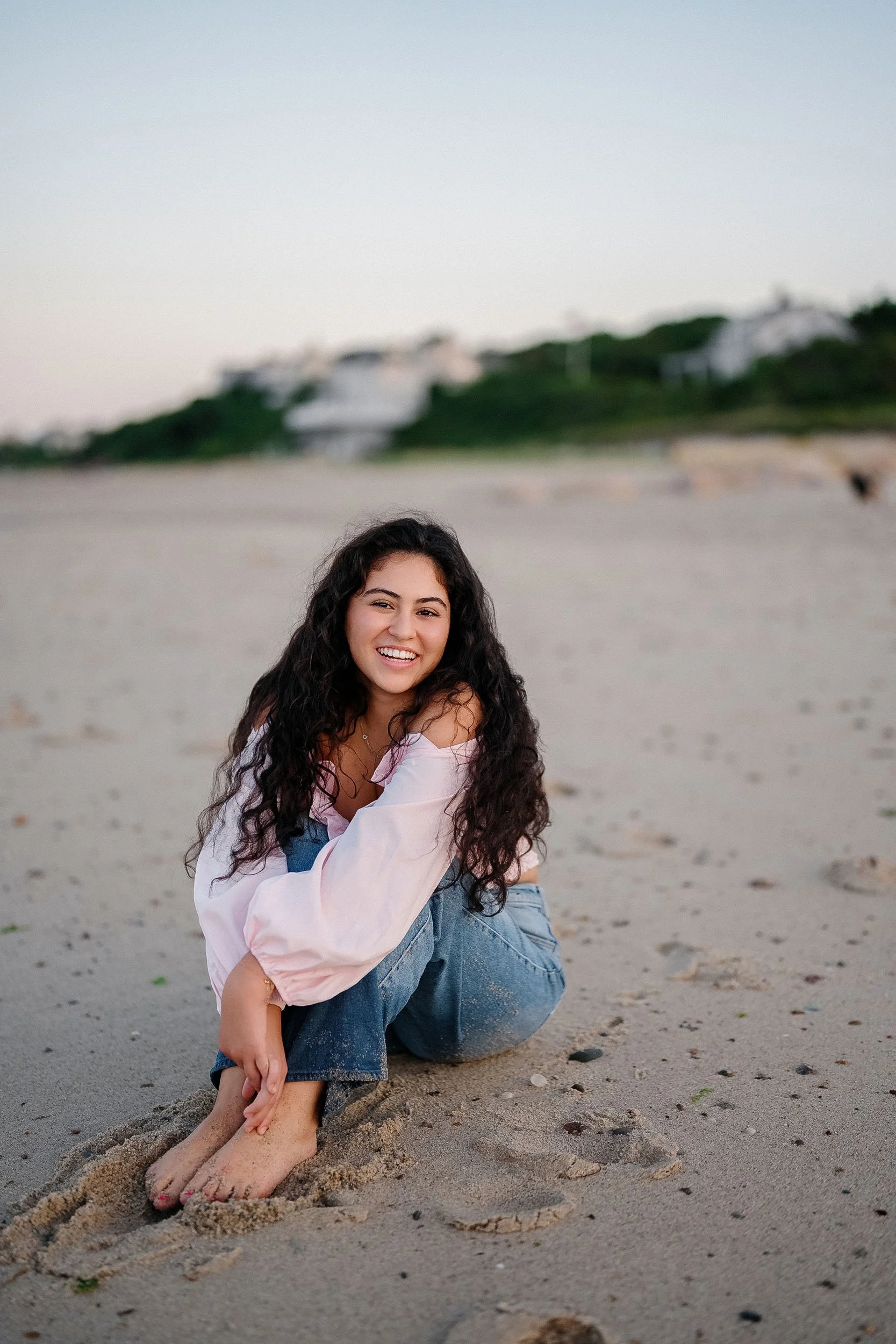 A young woman with long, curly dark hair, smiling and sitting on a sandy beach with her legs folded, wearing a light pink off-shoulder top and blue jeans.