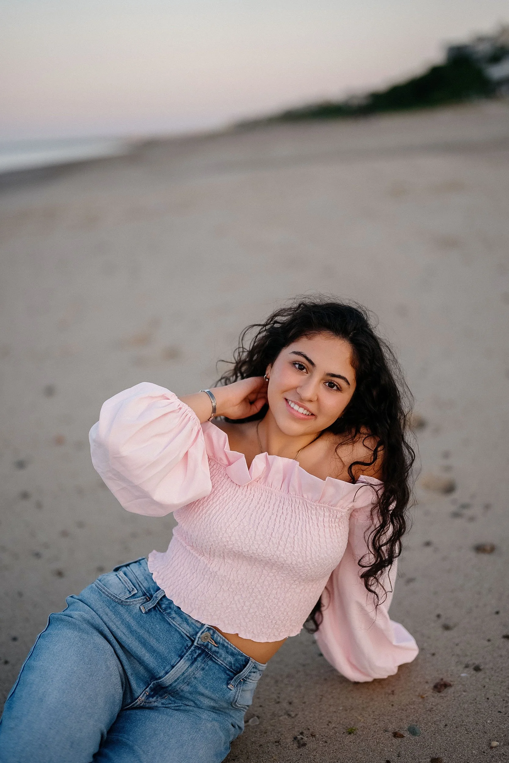 A young woman with curly dark hair sitting on the beach in the evening, wearing a pink off-the-shoulder top and blue jeans, smiling at the camera.