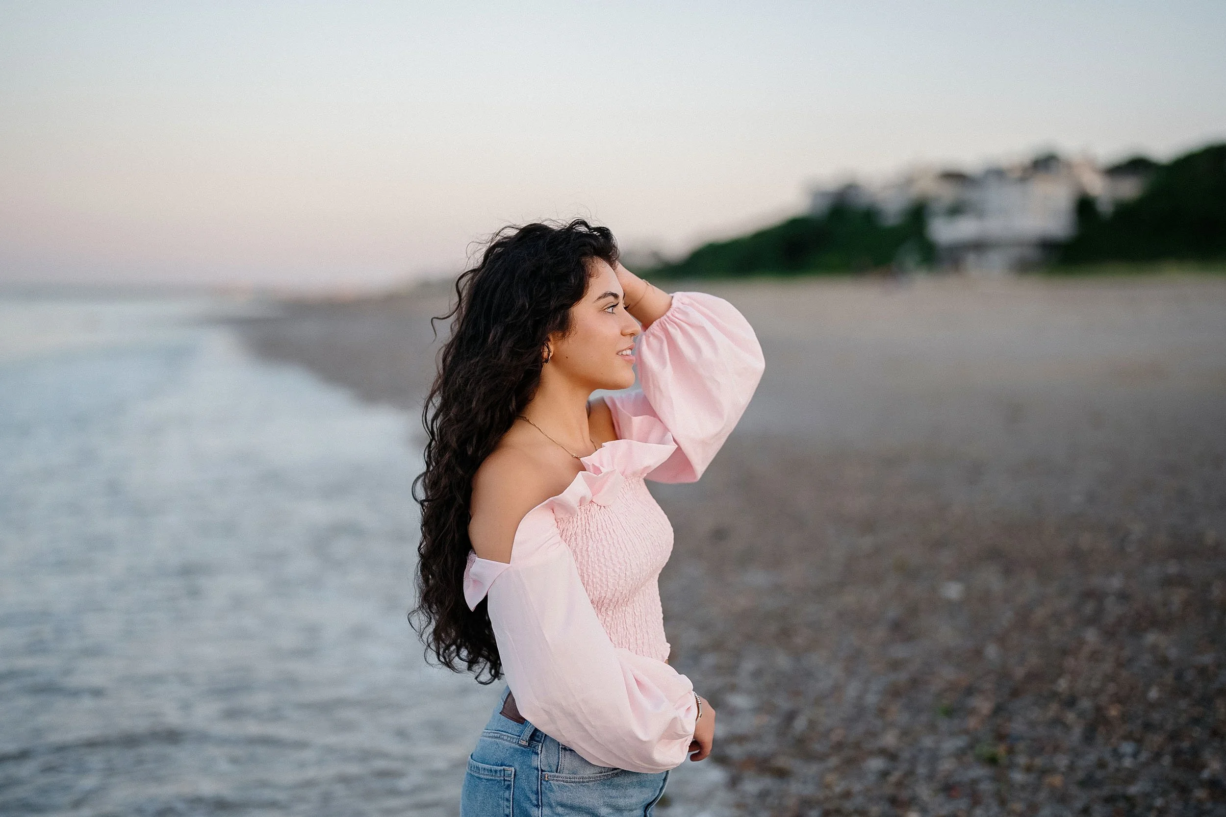 Young woman with long curly hair standing on a beach, looking towards the horizon during sunset or sunrise, wearing a pink off-shoulder top and jeans.