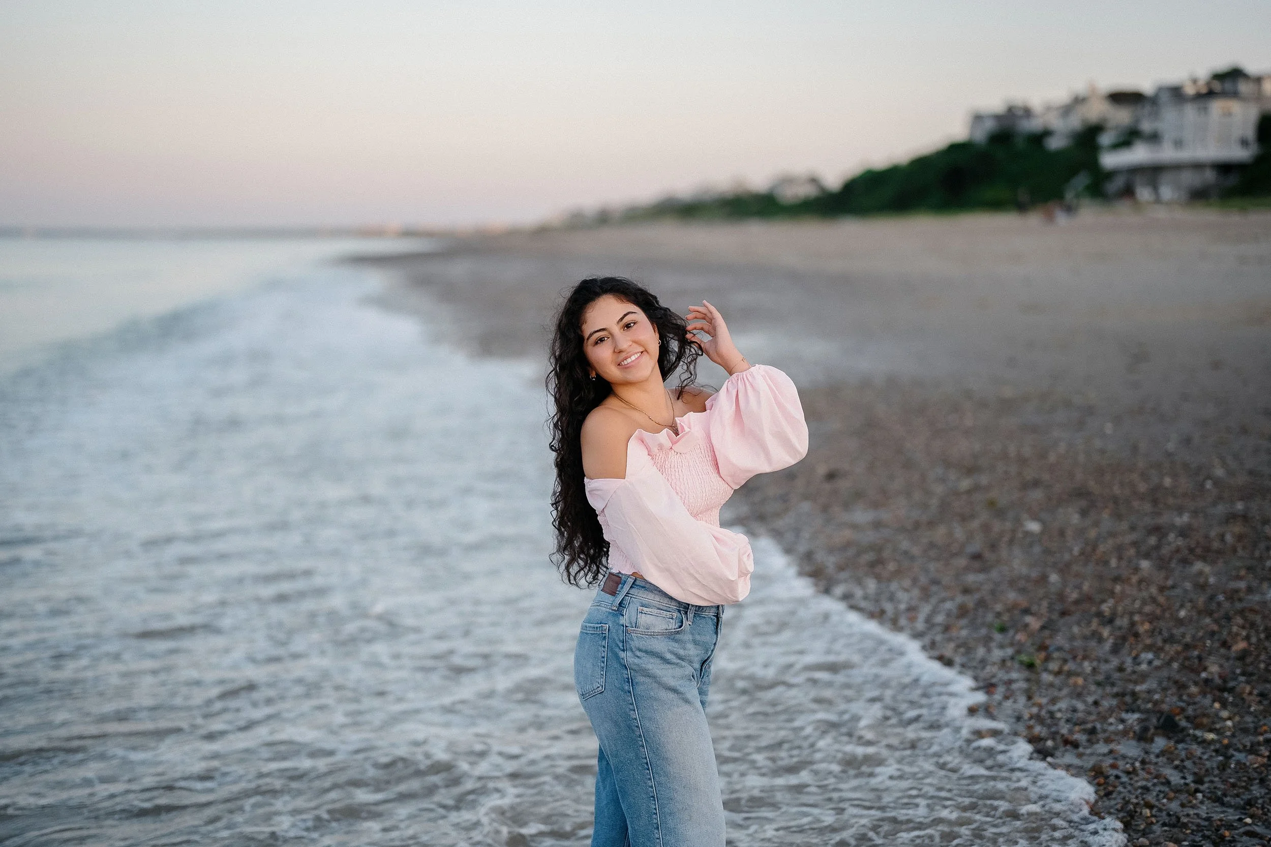 A young woman with long curly black hair smiling on a beach, wearing a pink off-shoulder blouse and blue jeans, with houses in the background and the ocean waves behind her.