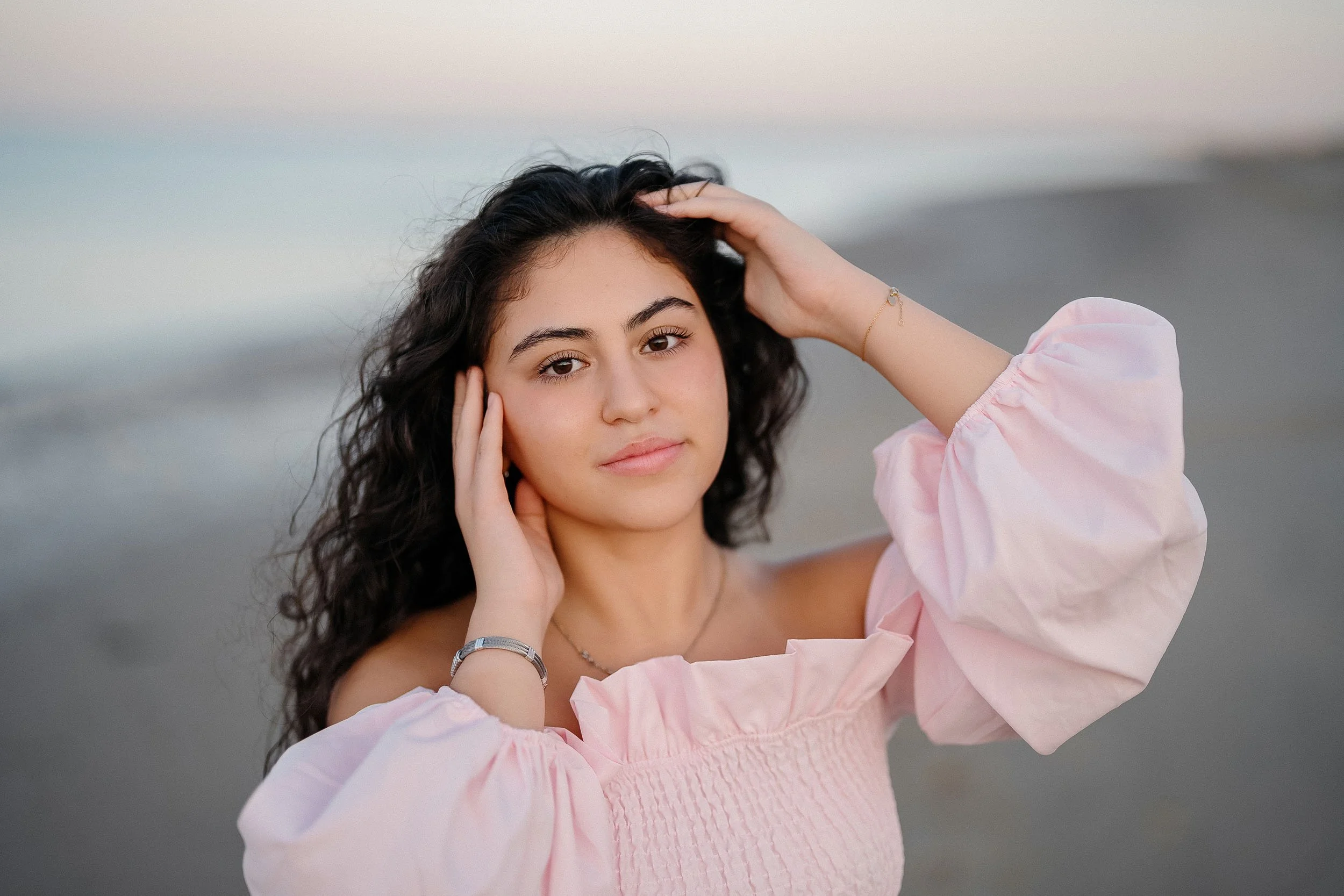 Young woman with dark curly hair standing on a beach, wearing a light pink off-shoulder top, holding her hair and facing the camera with a neutral expression.
