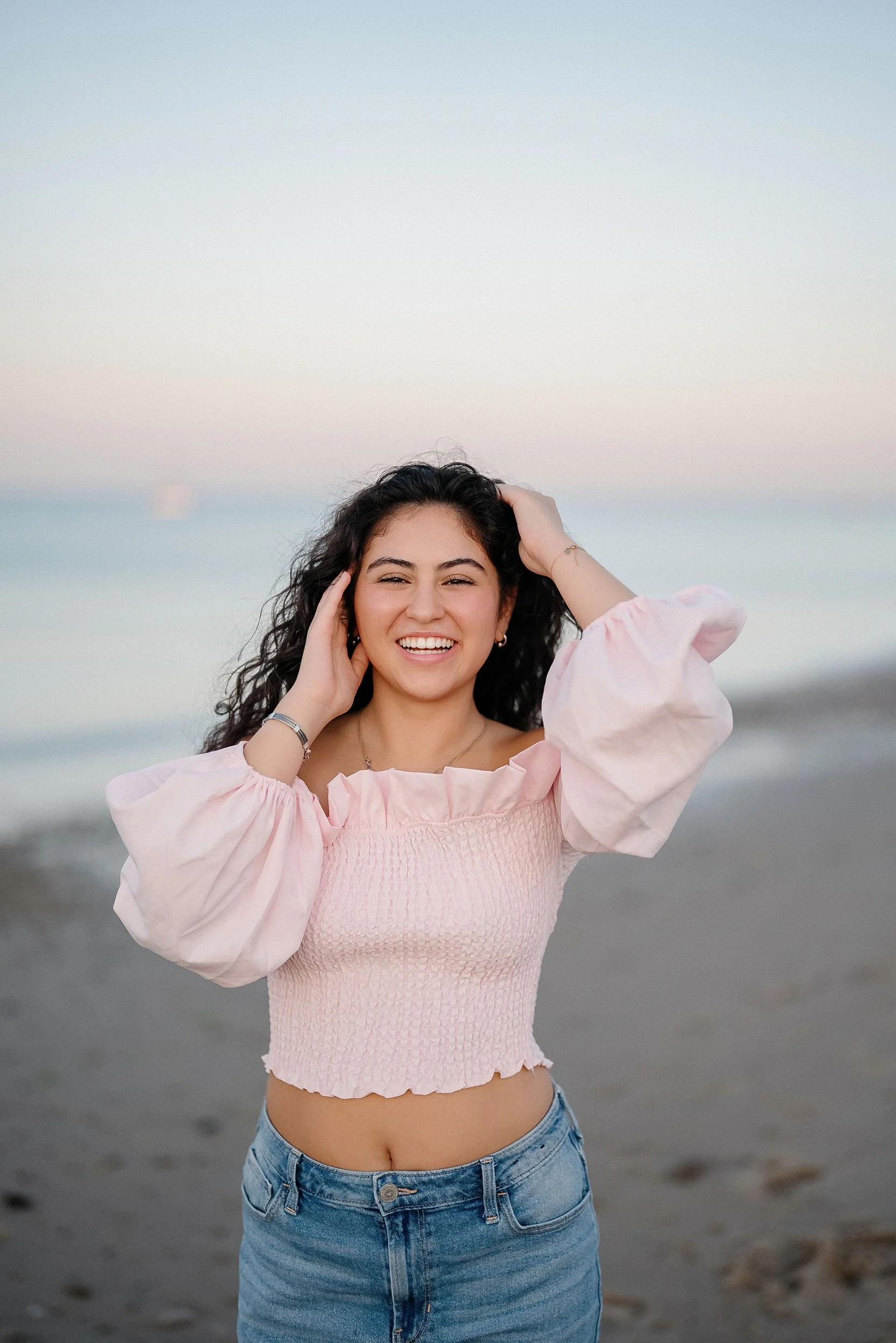 A young woman with dark curly hair, smiling, on a beach at sunset wearing a pink, puff-sleeve top and blue jeans.